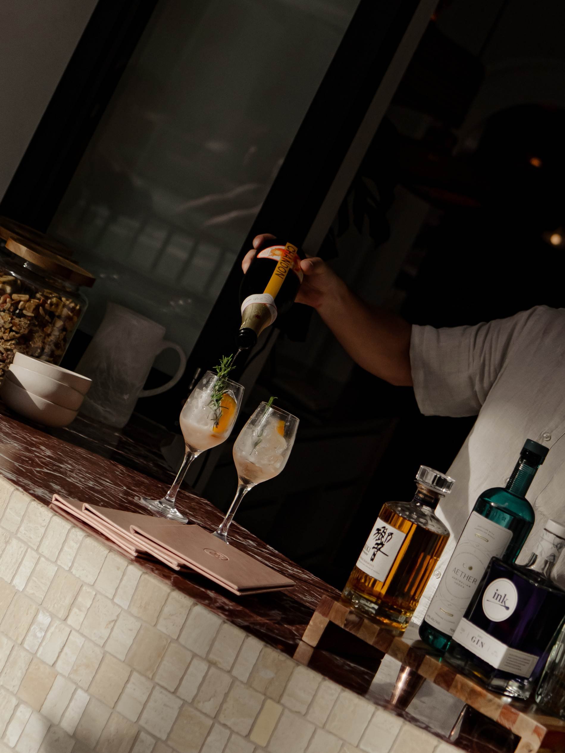 A person pouring liquor into two cocktail glasses with garnishes on a marble bar counter, with bottles of alcohol and glassware in the background. Featuring Basq House, a hotel in Byron Bay
