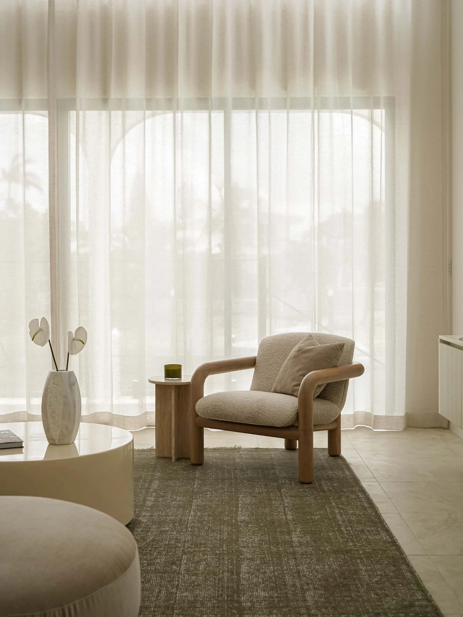 Bright living room with sheer curtains, a beige armchair with a cushion, a small side table with a green drink, a round coffee table with a vase and flowers, and a textured rug in luxury property in Mermaid Waters, QLD