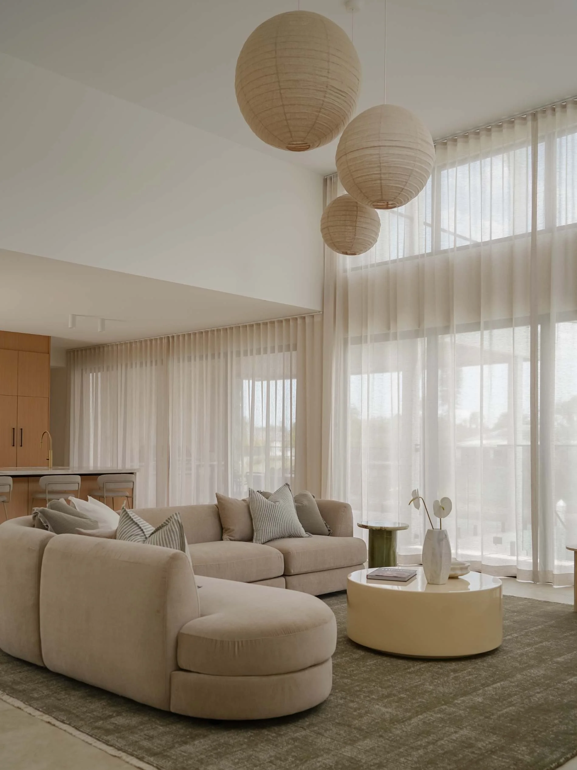 Living room with beige sectional sofa, natural light from large window with sheer curtains, modern decor, round coffee table with a vase, pendant light fixtures on ceiling in luxury Mermaid Waters, QLD 