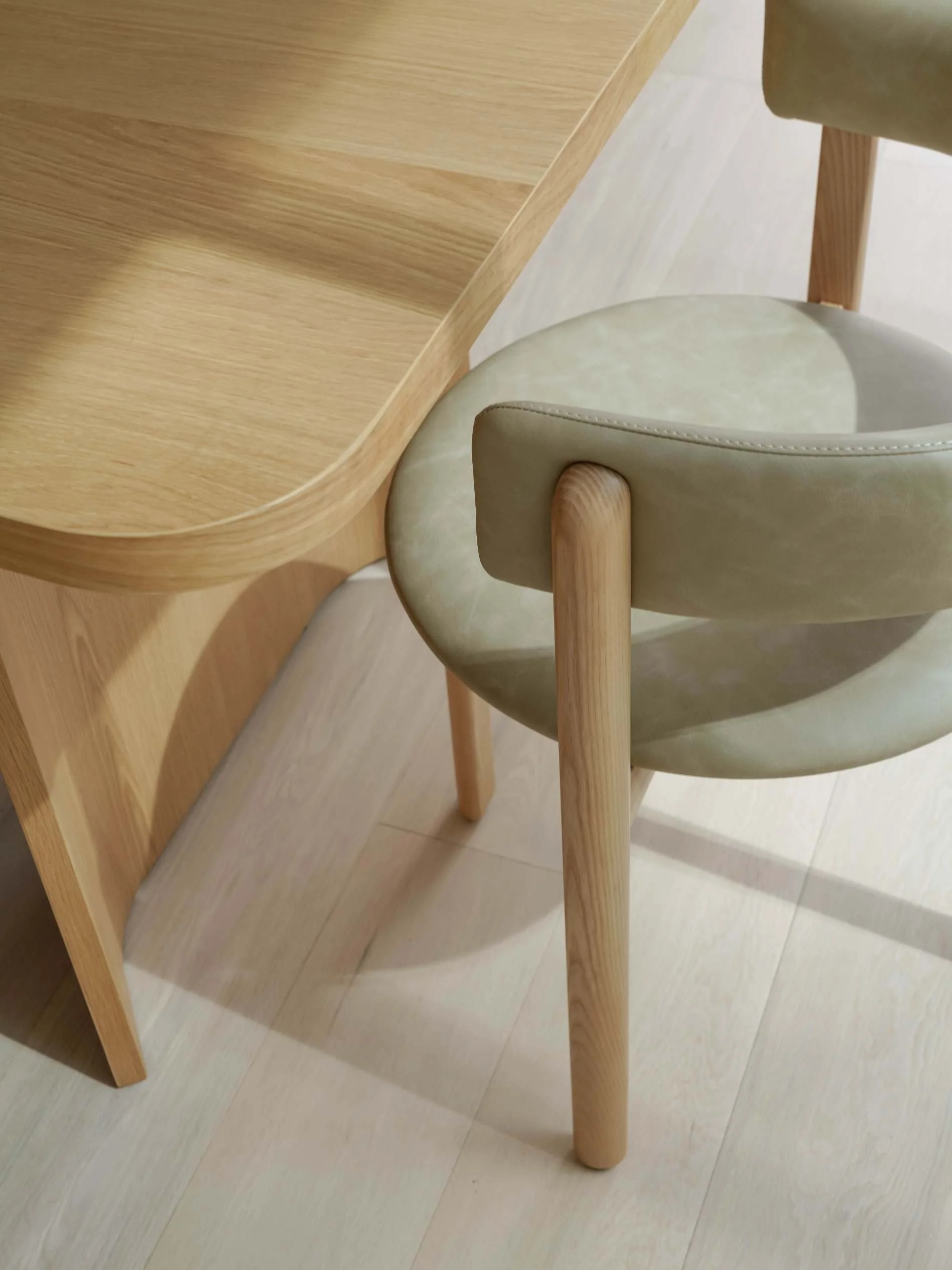 Close-up of a wooden chair with a light green cushion, positioned next to a wooden table with rounded edges, on a light-colored wooden floor in Ostani House 