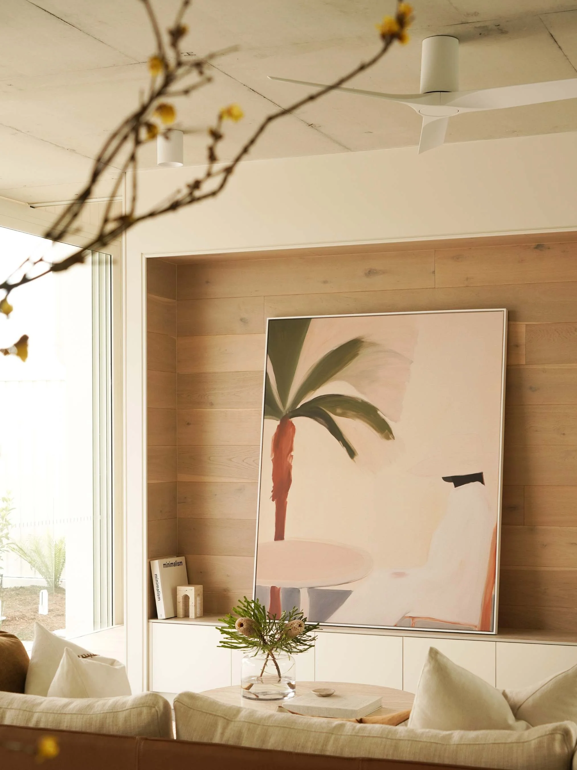Modern living room with a large painting of a palm tree on a wooden wall, a low white sideboard, and a glass vase with greenery on a table in front of a beige sofa in Noosa Heads, QLD