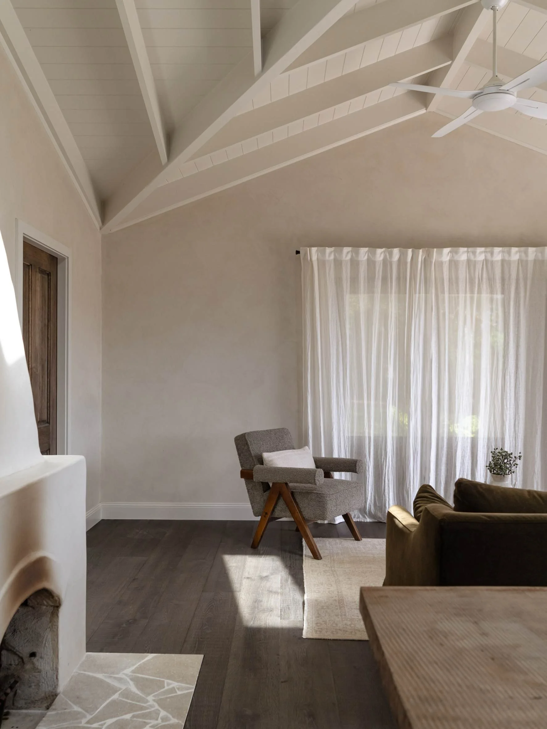A cozy living room corner with a gray armchair, a brown sofa, and a small rug. There is a large window with sheer white curtains, a white ceiling fan, and exposed ceiling beams at Hunter Cabin in Byron Bay