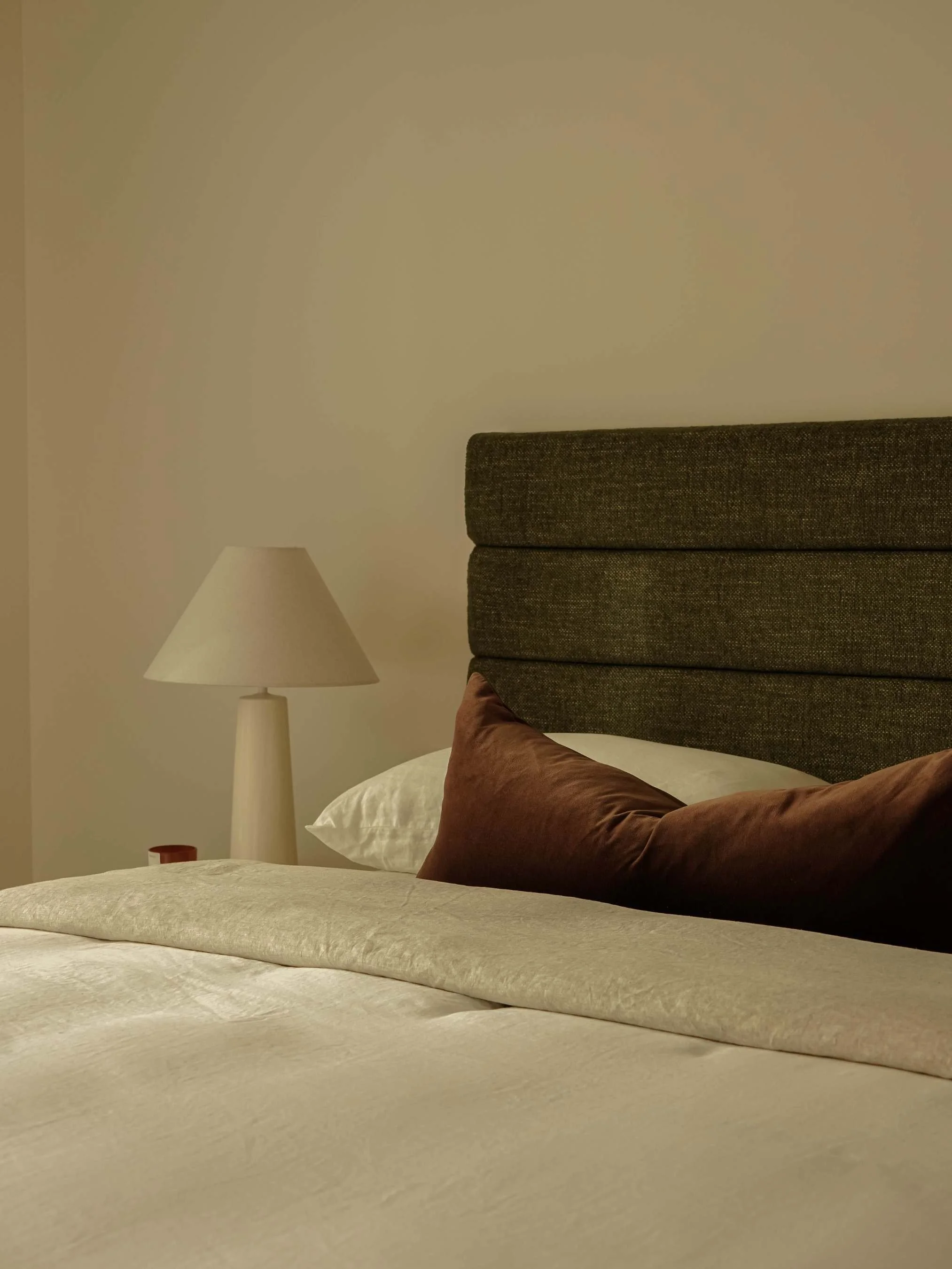 A bedroom corner with a bed featuring a dark upholstered headboard, a brown pillow, a white pillow, a beige bedspread, a white table lamp with a beige shade, and a small red object on the side.