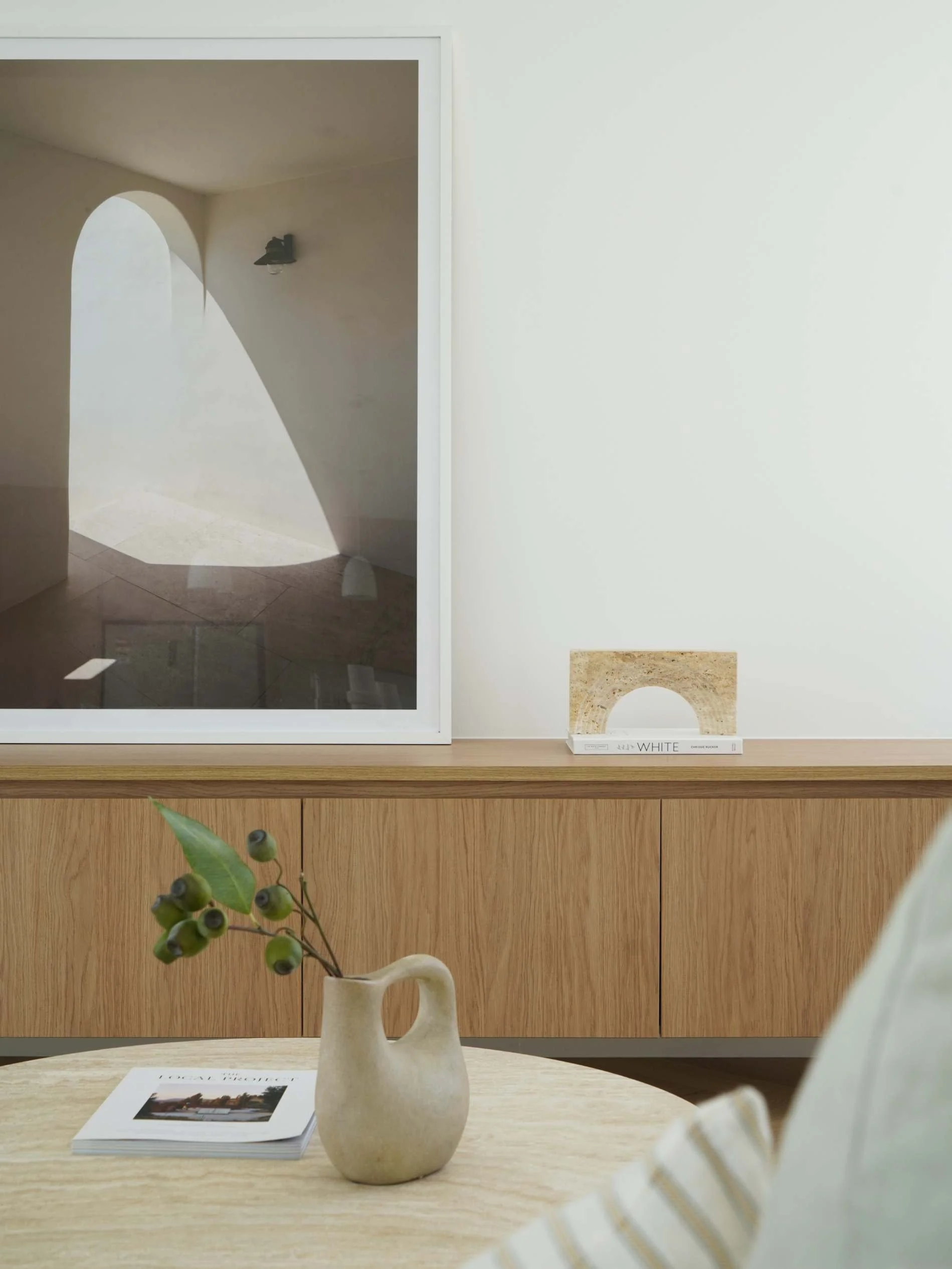 A minimalist interior with a wooden sideboard, a ceramic vase with green plant stems, a white book titled 'WHITE' with a marble-like object on top, and a blurred chair with striped cushion in the foreground. A framed photograph leans against the wall