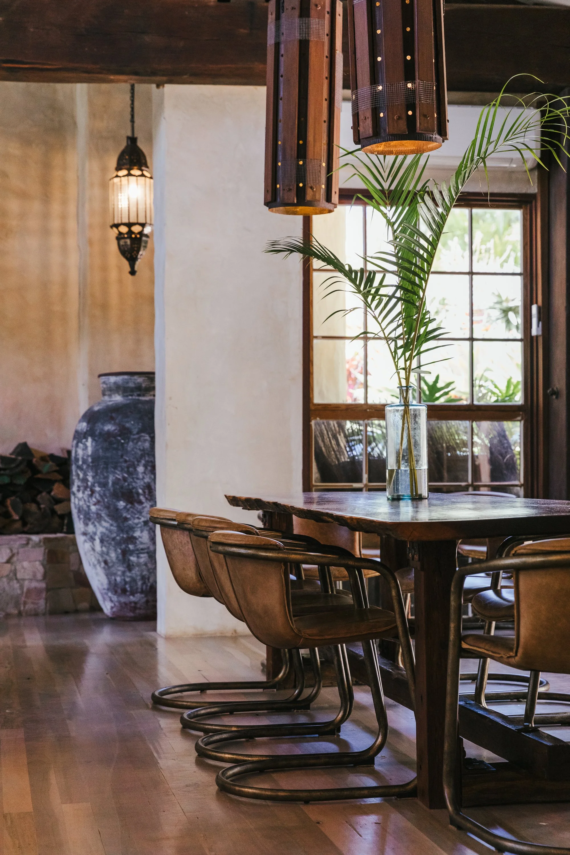 Interior of a rustic dining room with a wooden table, leather chairs, a large potted plant, wooden pendant lights, and a window with sunlight streaming through.