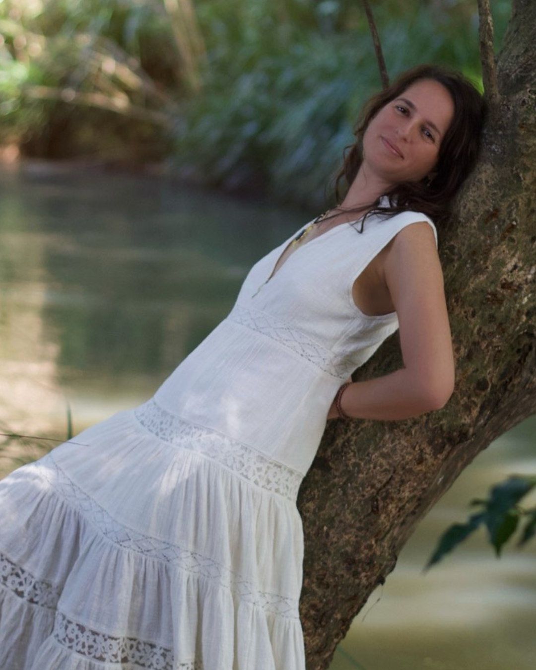A young woman in a white dress leaning against a tree near a body of water, smiling softly.