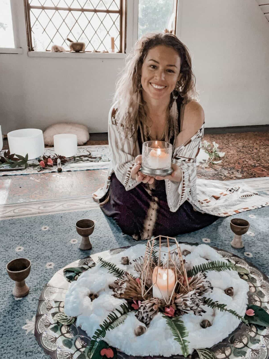 A woman with curly hair sitting on the floor, holding a candle in a glass holder, smiling at the camera. She is surrounded by a decorative setup with a candle inside a geometric frame, greenery, and flowers on a white cloth, with small wooden cups around her. A window with a lattice design and some bowls is visible in the background.