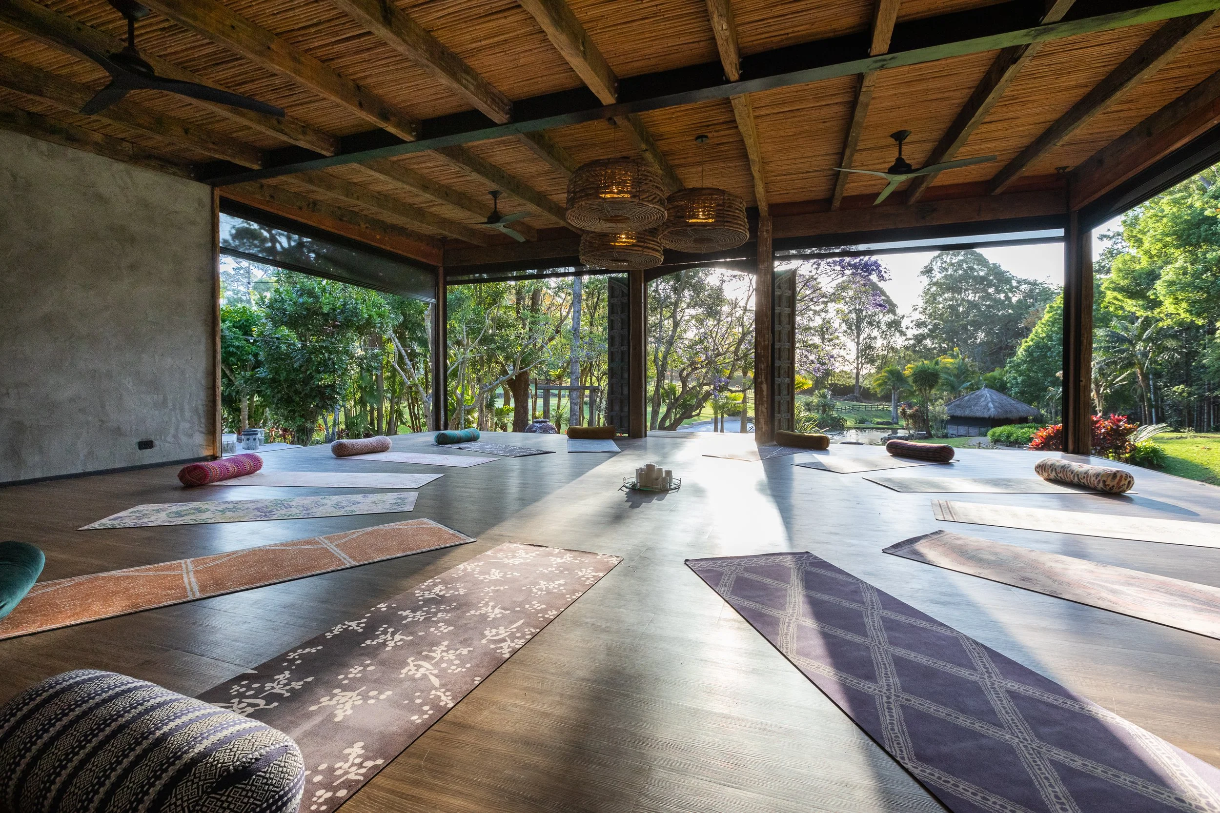 Indoor yoga studio with large open windows, wooden ceiling, and yoga mats and bolsters arranged on the floor, overlooking a lush green outdoor landscape.