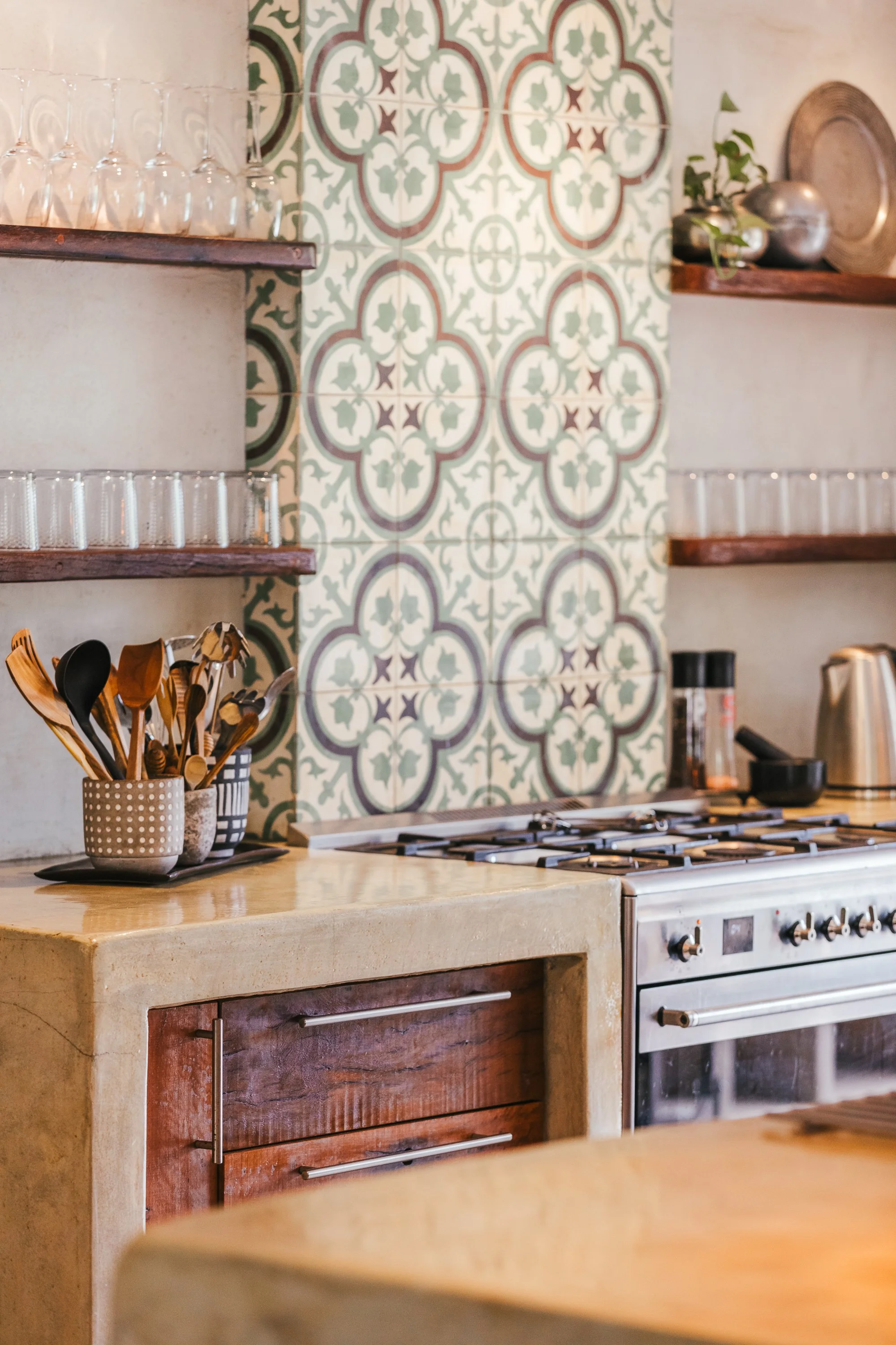 A cozy kitchen with a decorative tiled backsplash, open wooden shelves holding glassware and decorative items, a countertop with cooking utensils in cups, a stove, and various small appliances.