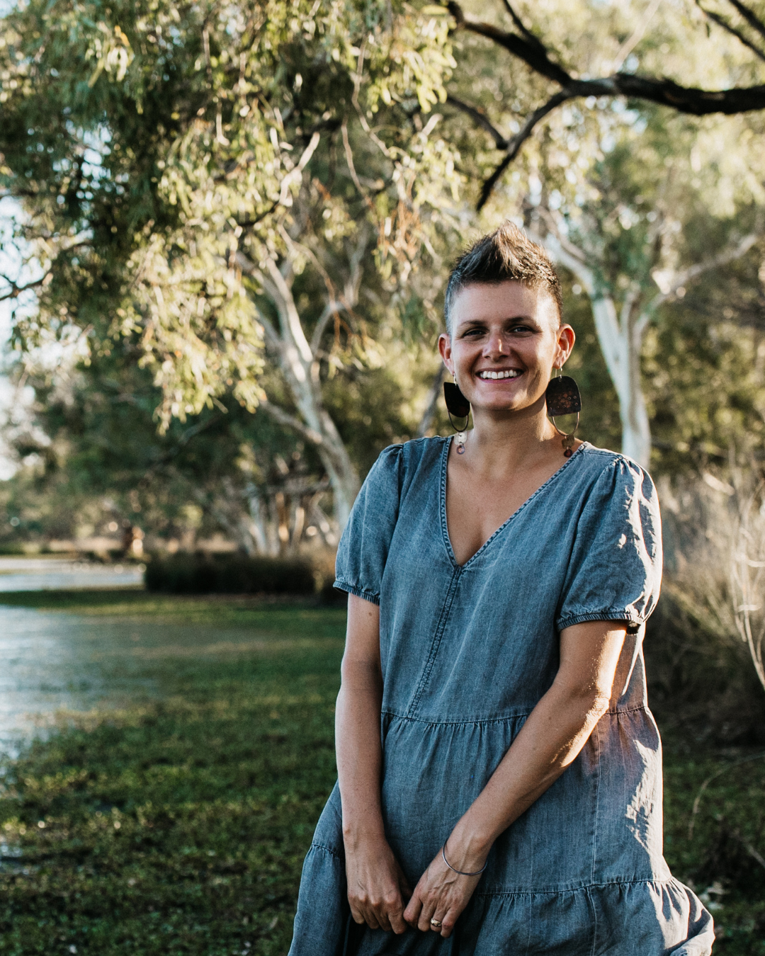 A smiling woman with short hair wearing a denim dress and large earrings, standing outdoors in a park with trees and grass.