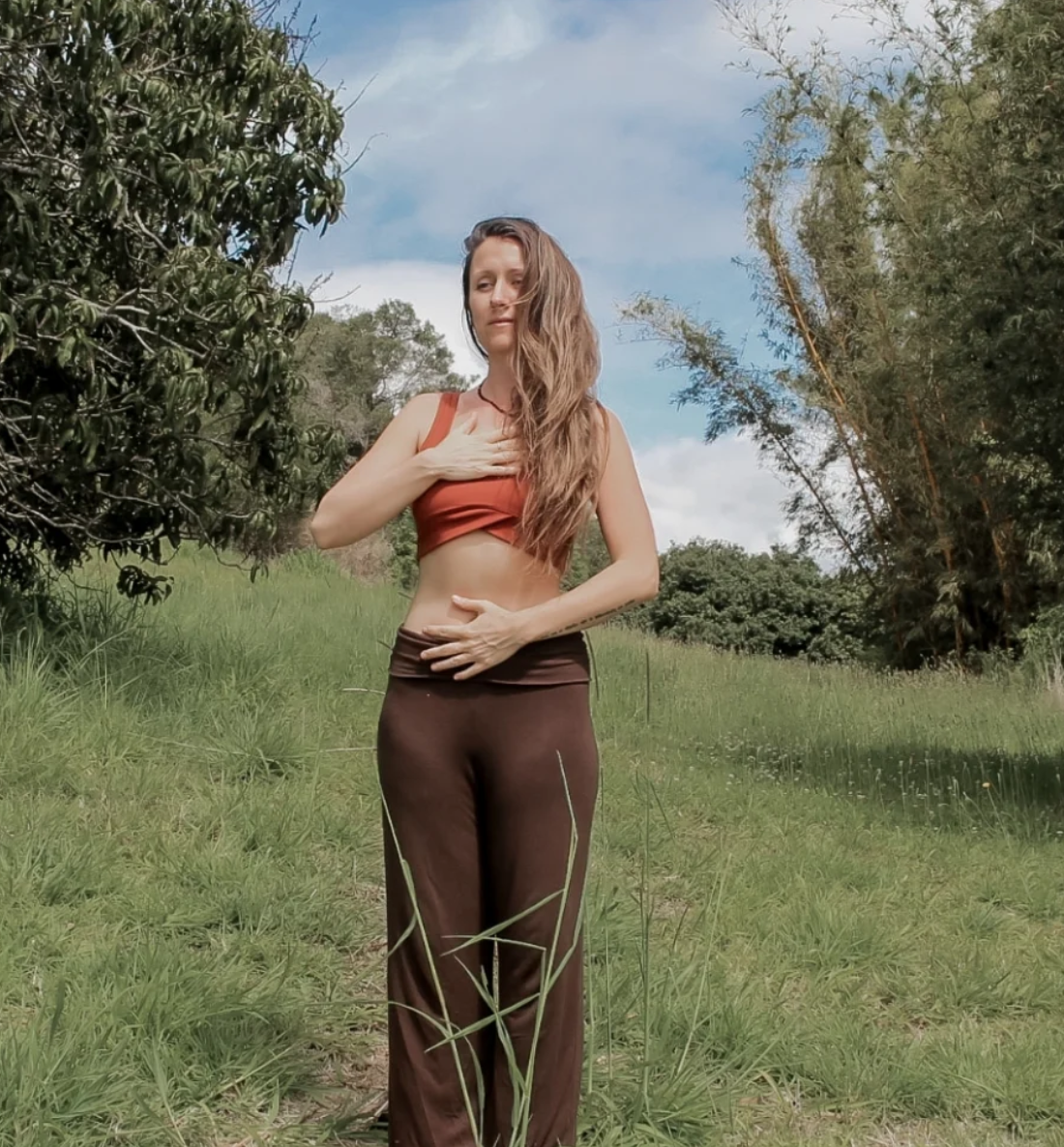 A woman with long hair, wearing a red crop top and brown pants, stands outdoors in a grassy area surrounded by trees, holding her hands over her chest and stomach, with a blue sky and clouds overhead.