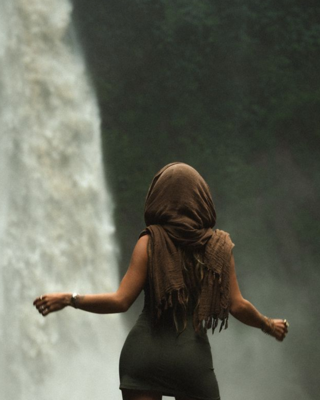 A woman wearing a headscarf and a black dress walking near a waterfall in a lush green forest.