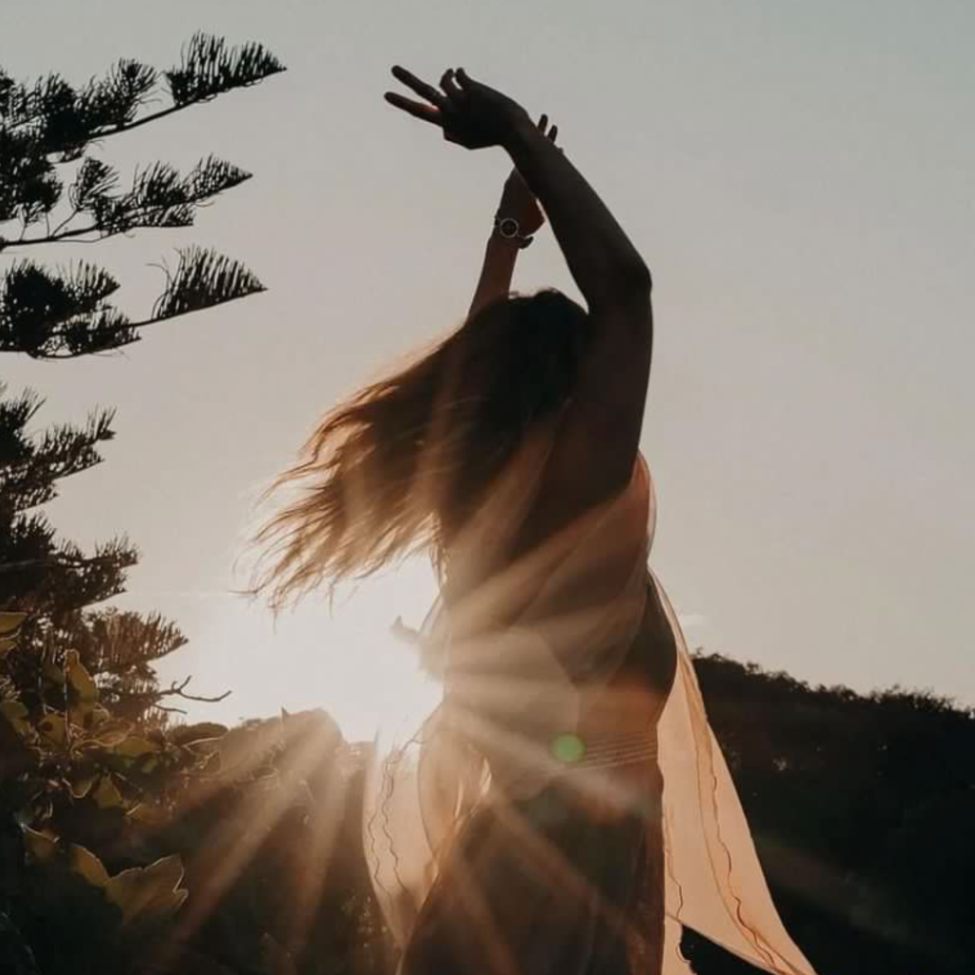 A woman with long hair dancing outdoors at sunset, with her arms raised above her head and sunlight streaming through her, surrounded by trees and foliage.
