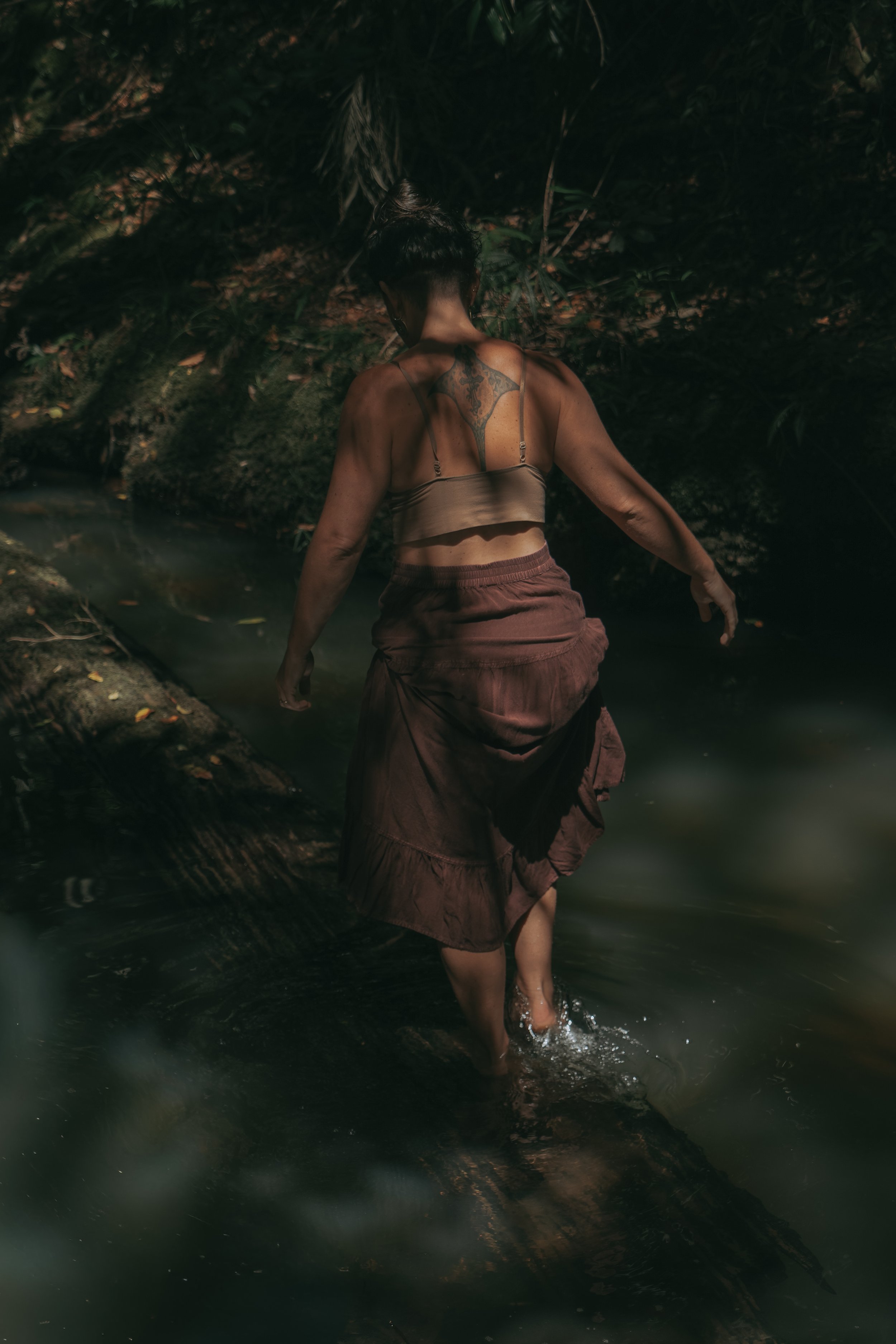 A woman walking through a shallow stream in a forest at night, wearing a crop top and a long skirt, with her back to the camera.
