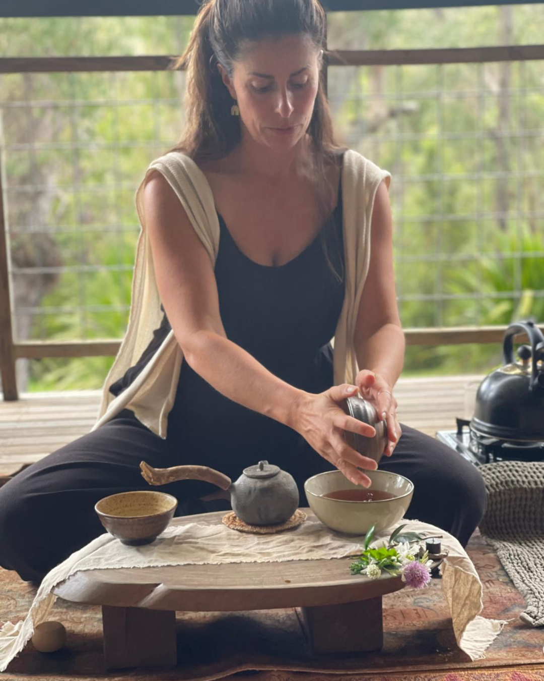 Woman sitting on a wooden porch, pouring tea into a bowl, surrounded by tea utensils and flowers, with greenery in the background.