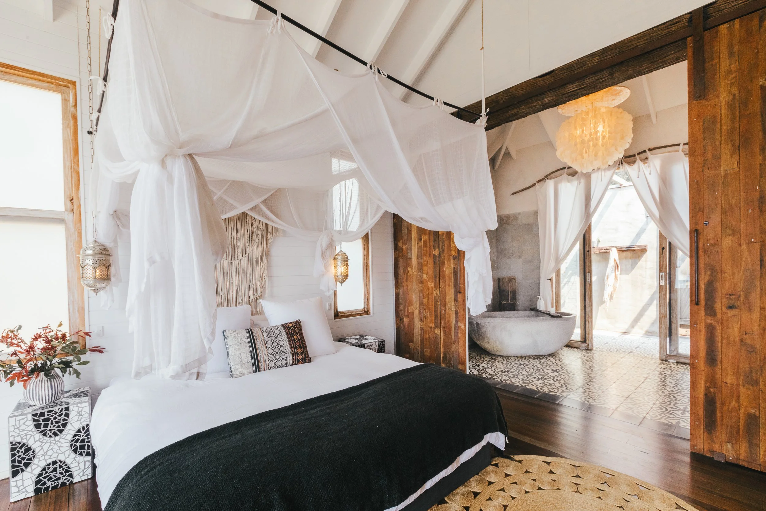 A cozy bedroom with a white bed featuring a black blanket, decorated with patterned pillows, hanging white curtains from a canopy, and warm lighting from lantern-style fixtures. An open sliding door leads to a bathroom with a freestanding bathtub, and the room features wood accents and natural light.