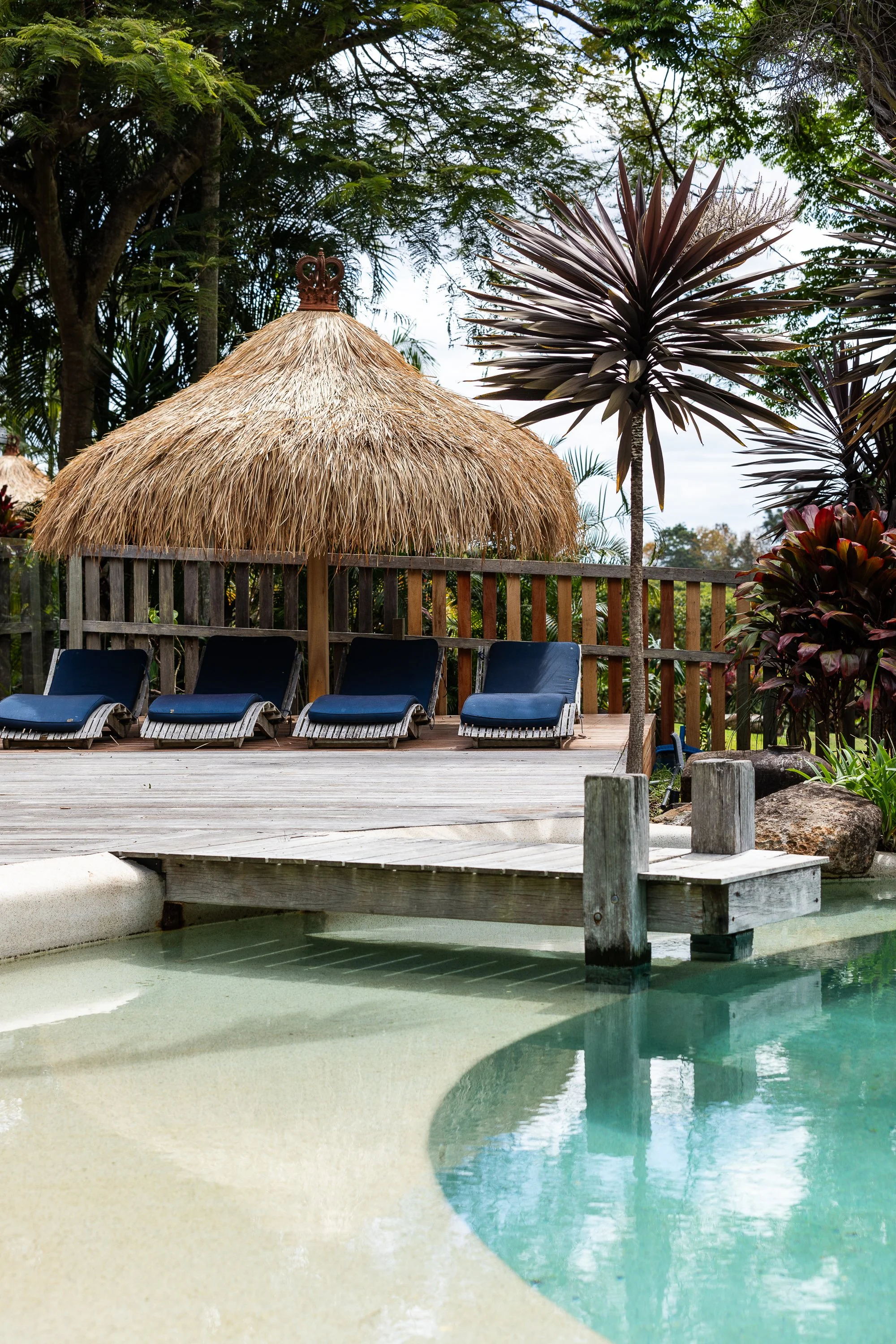 Poolside area with several black lounge chairs under a thatched-roof tiki hut, tropical plants, and a wooden dock over the water, with lush green trees in the background.