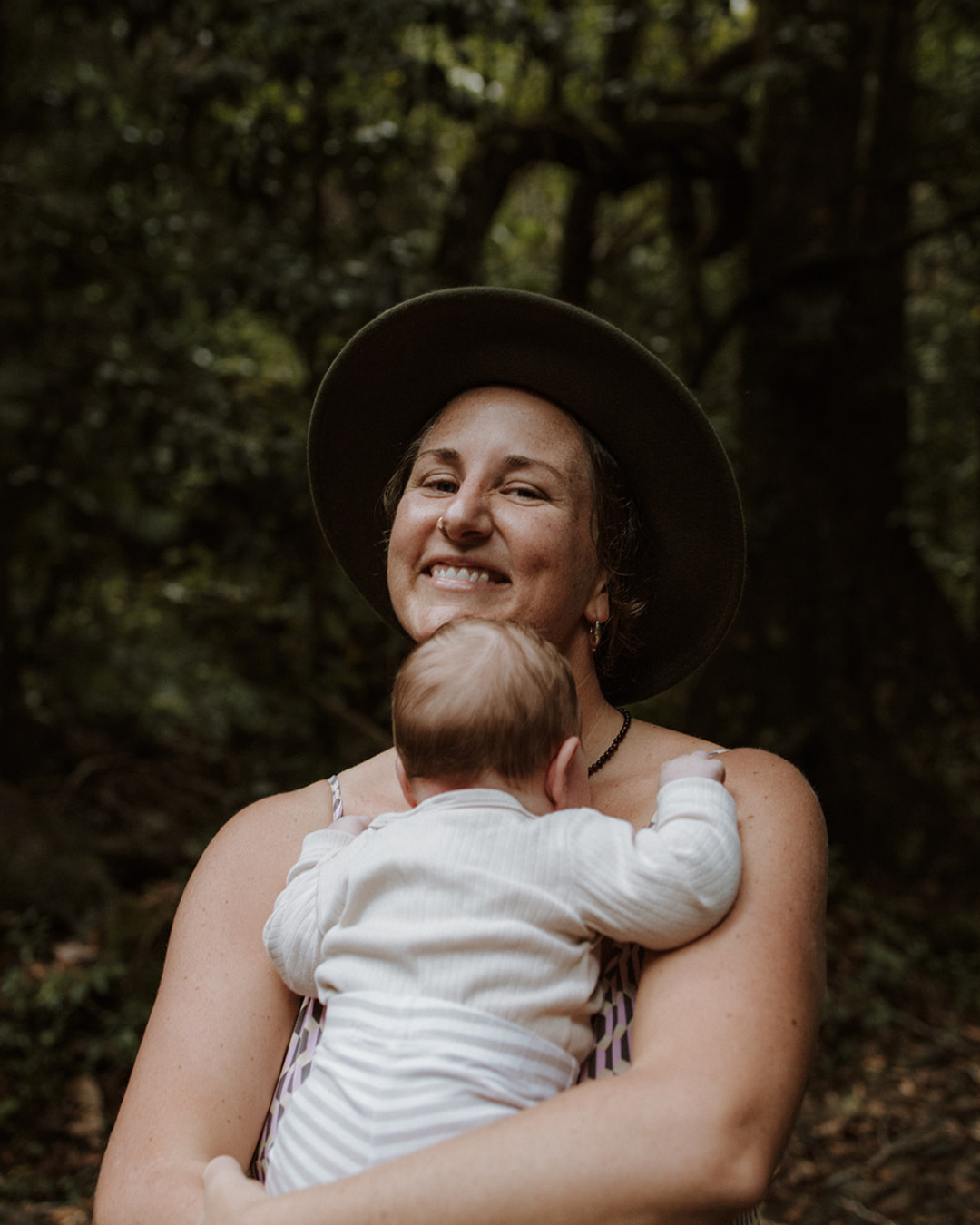 A woman wearing a wide-brimmed hat holding a baby in an outdoor wooded area.