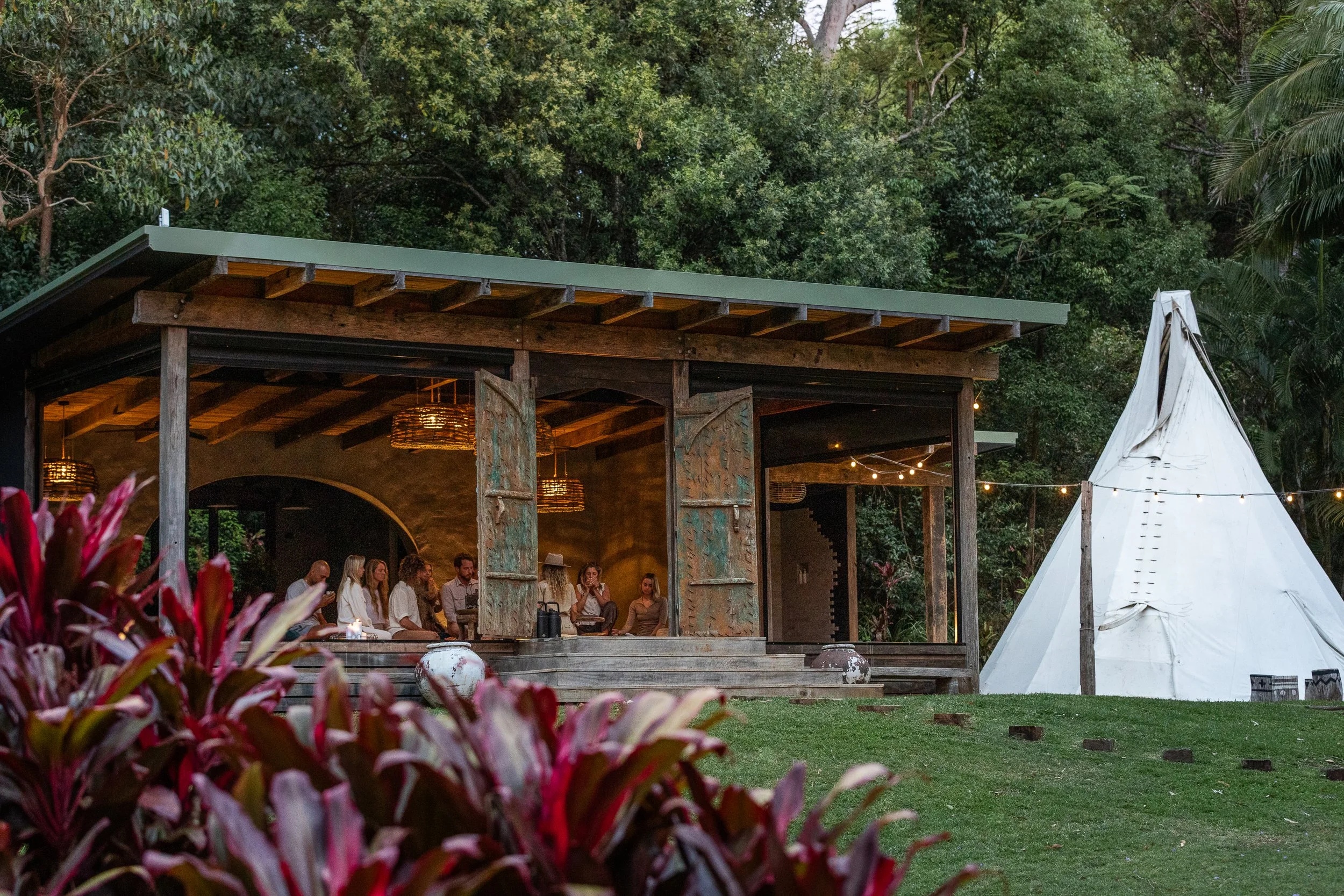 People gathering inside an open wooden structure with a green roof, adjacent to a white teepee with string lights, surrounded by lush greenery and trees.
