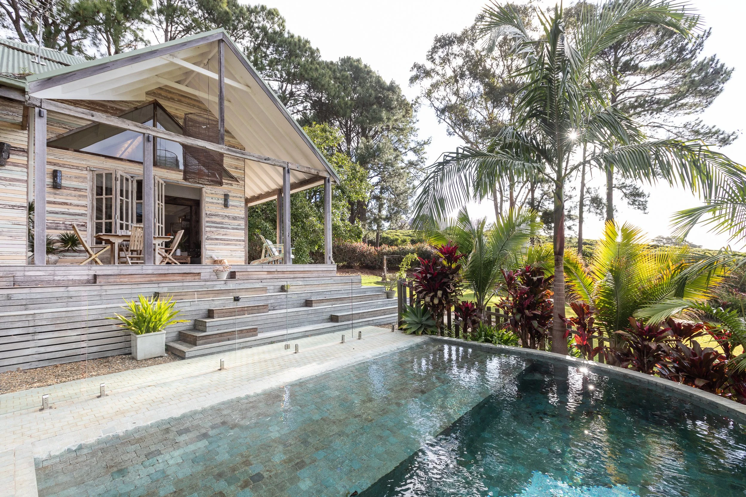 A wooden house with a patio overlooking a small pool surrounded by tropical plants, including tall palm trees.
