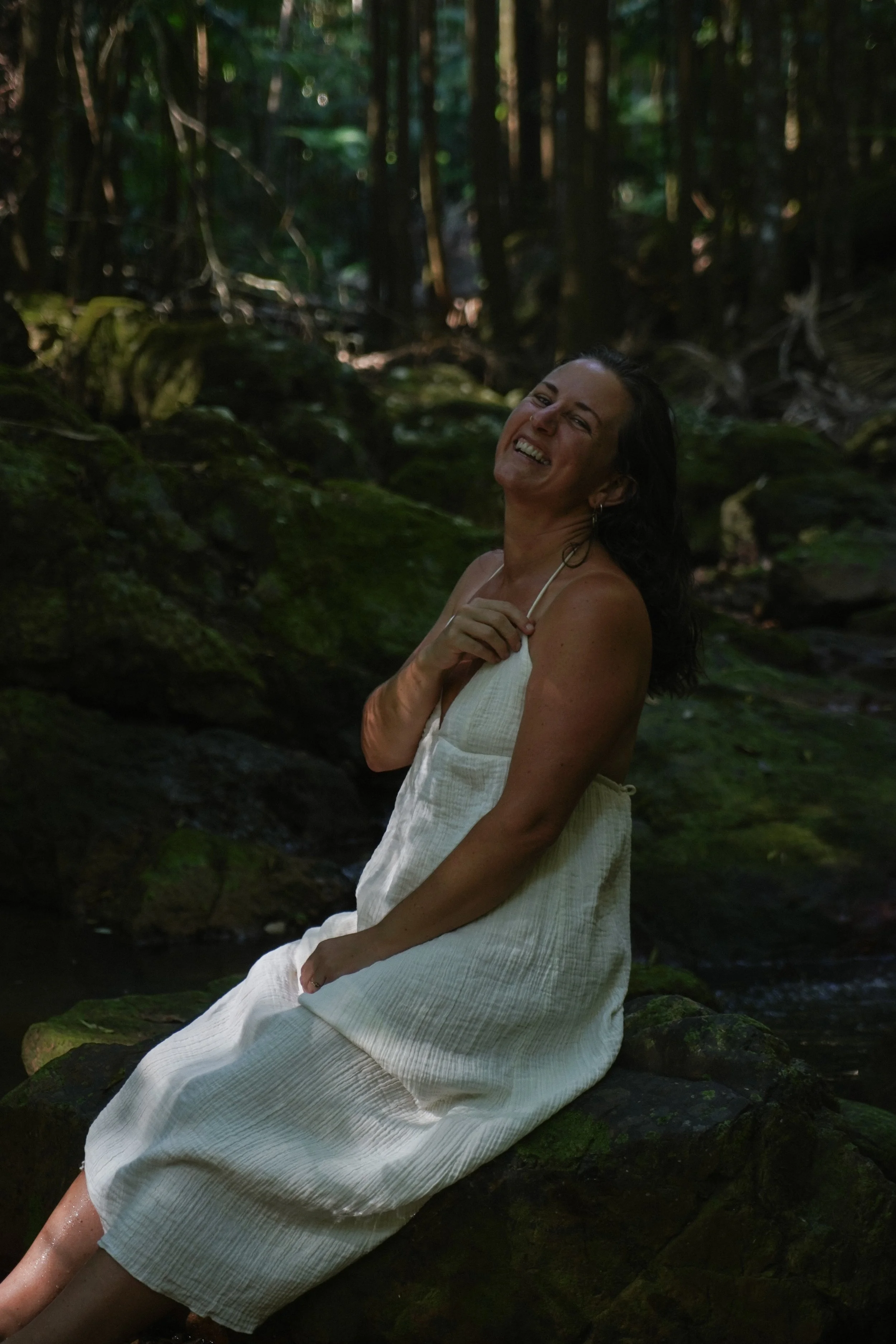 A woman in a white dress sitting on moss-covered rocks in a lush green forest, smiling and enjoying the natural scenery.