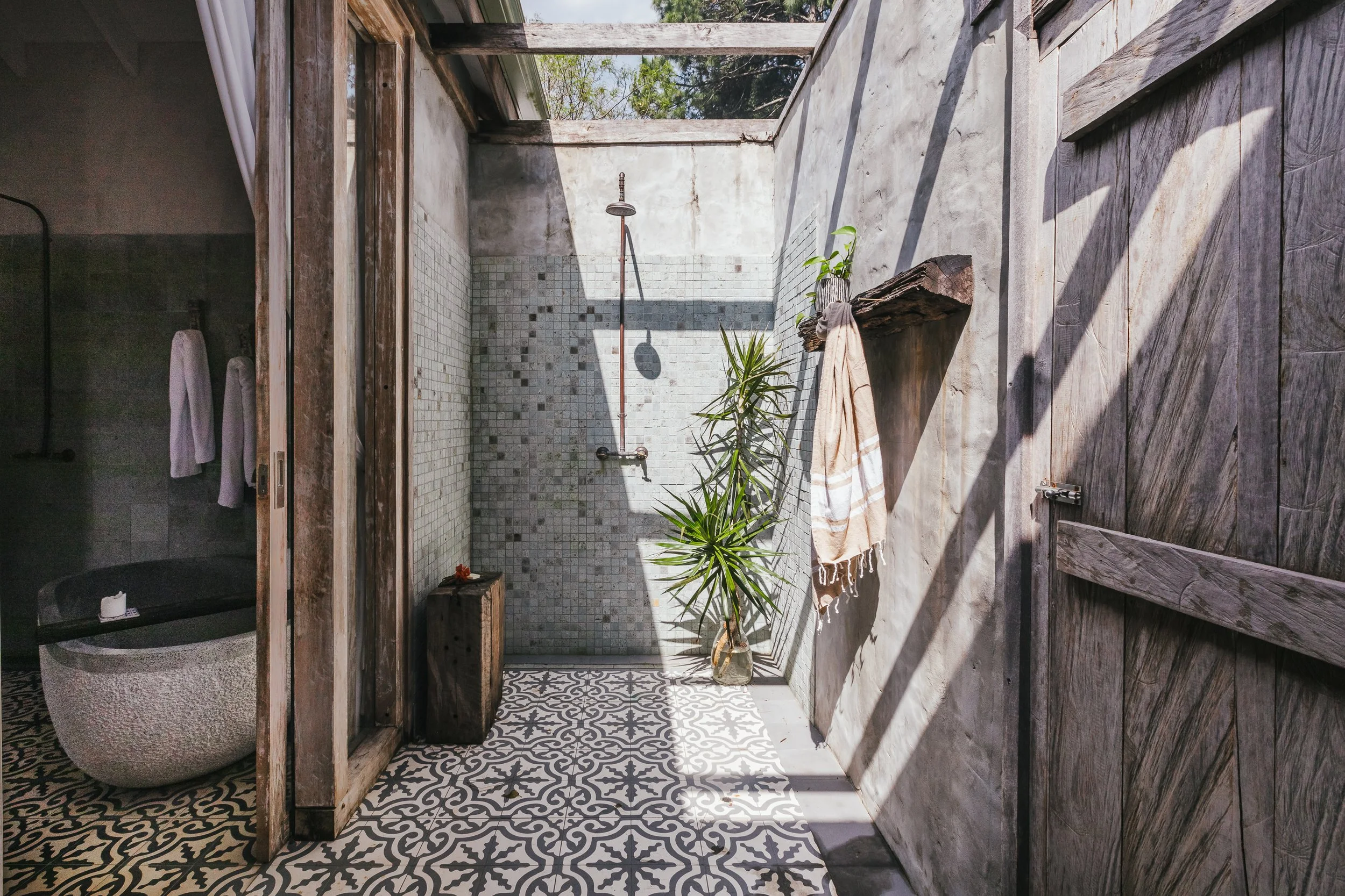 Outdoor shower area with patterned tile floor, a potted plant, and a wooden shelf, partially enclosed by wooden and concrete walls, with sunlight casting shadows.