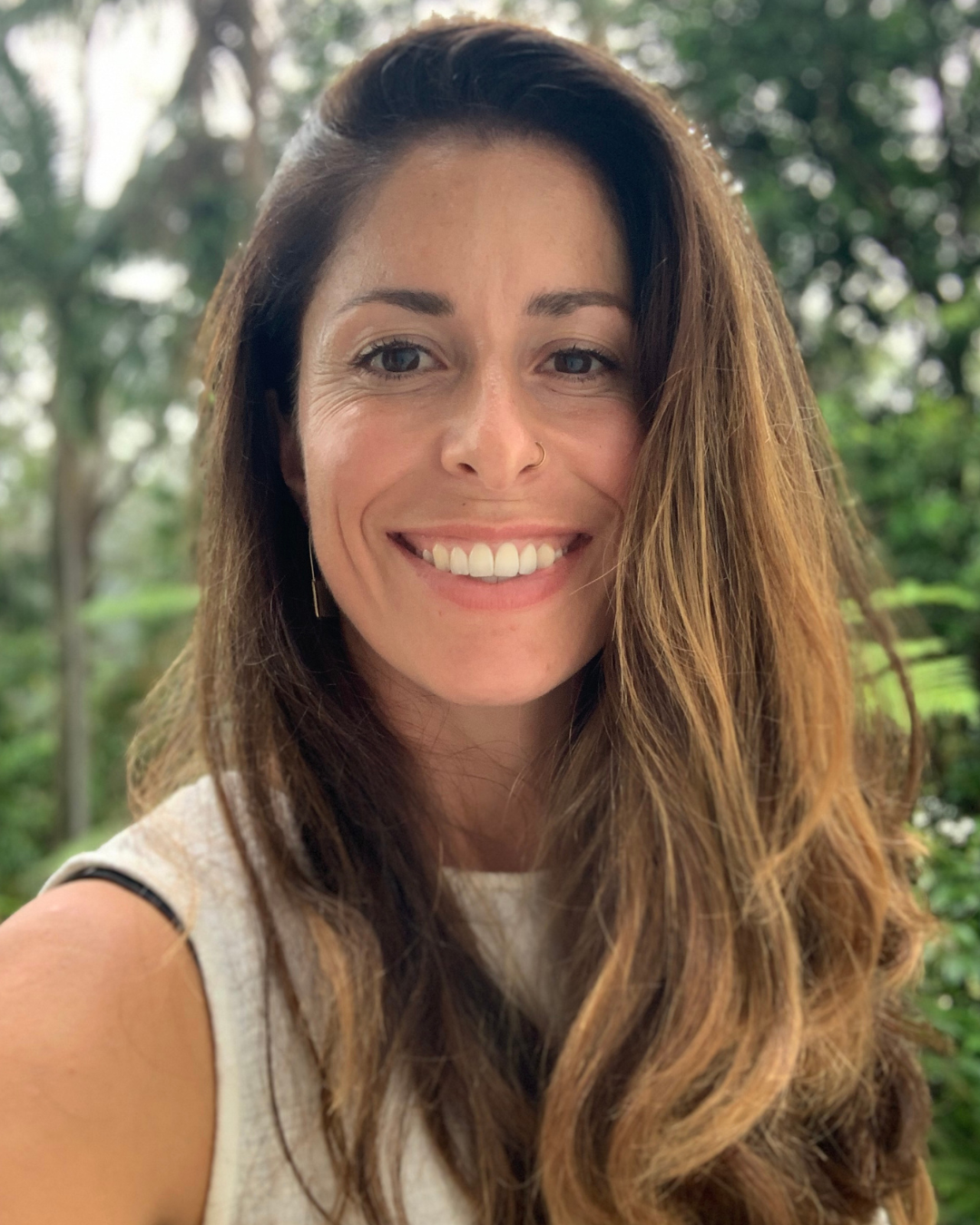 A smiling woman with long brown hair, wearing a white sleeveless top, standing outdoors with greenery in the background.