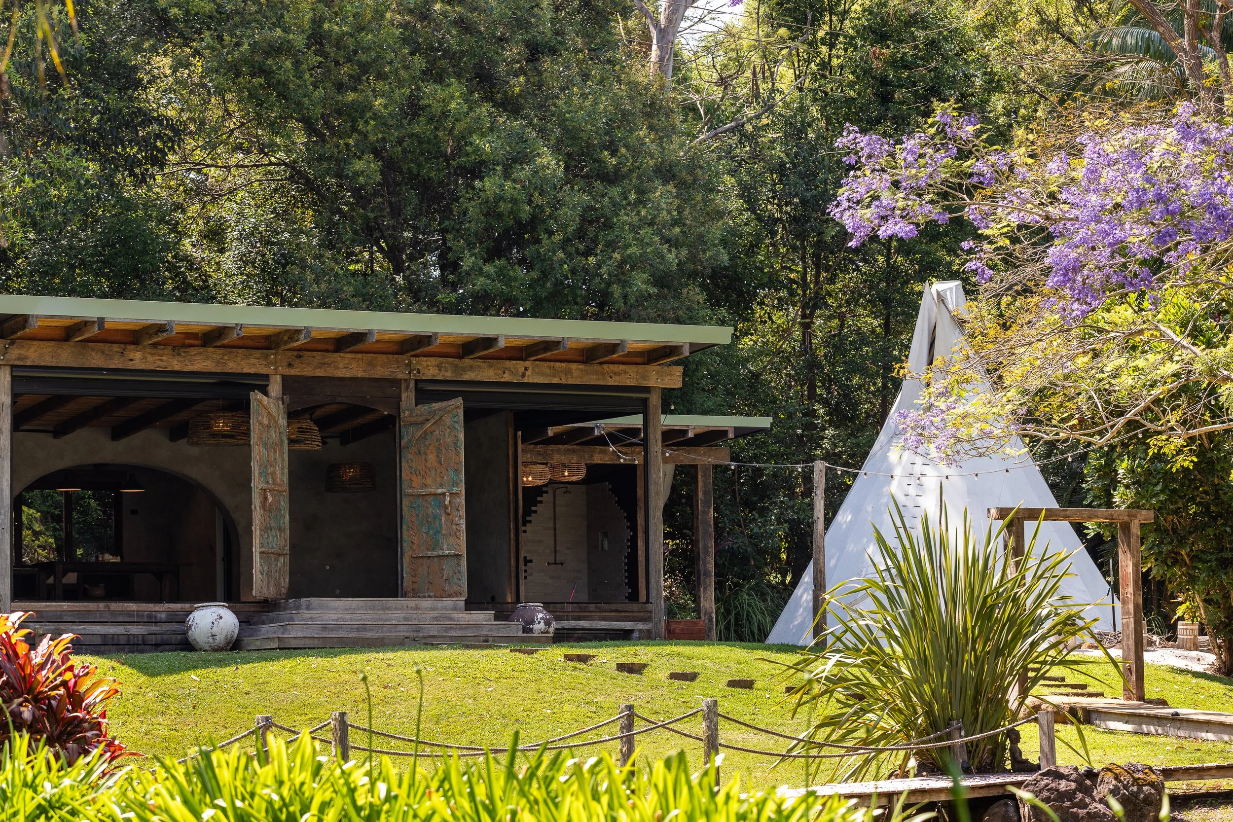 A rustic outdoor scene with a small modern building, a teepee tent, and lush greenery and flowering trees in the background.