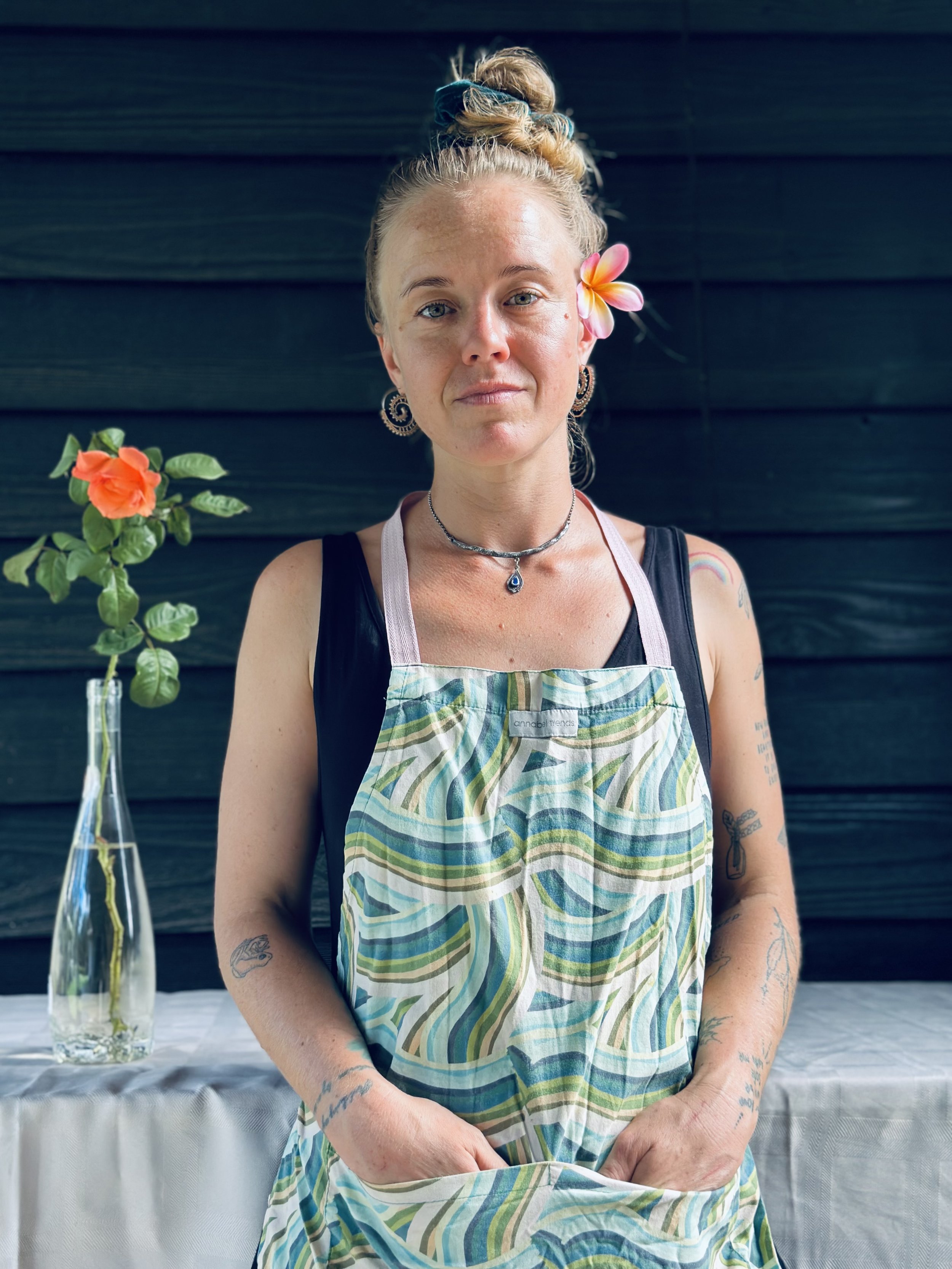A woman with blonde hair tied up, decorated with a flower, standing in front of a dark wooden wall. She is wearing a colorful apron over black clothing, has tattoos on her arms, and is looking at the camera. There is a table with a white tablecloth and a vase with pink and orange flowers on her left.