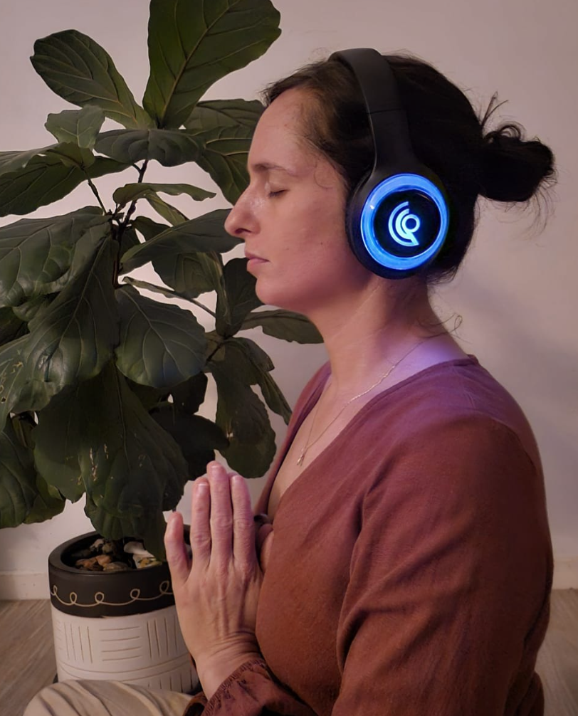 A woman with dark hair in a bun, wearing headphones and a brown top, practicing meditation with her hands in a prayer position beside a large green houseplant indoors.