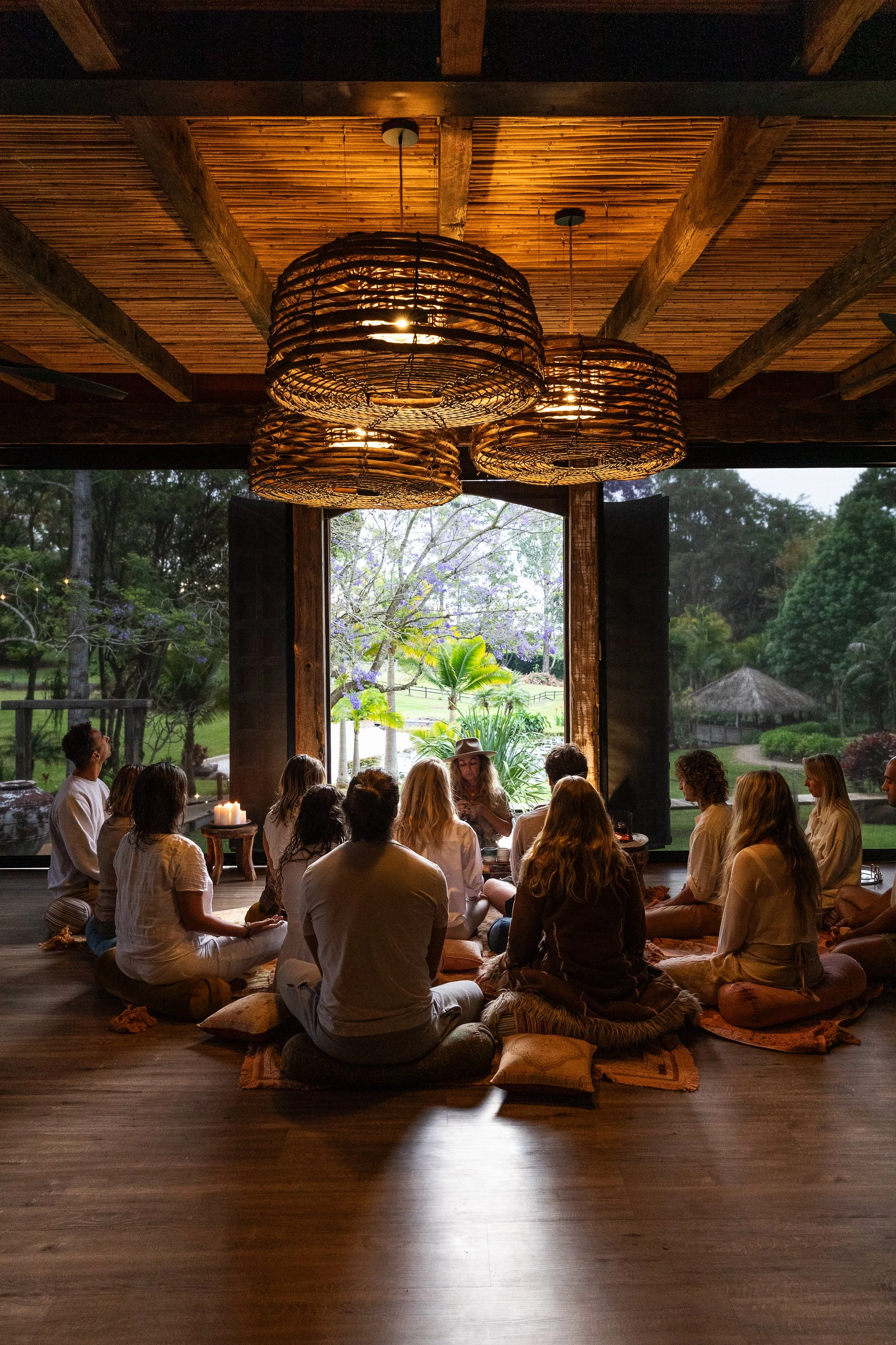 Group of people sitting on cushions and meditating indoors, with large open window showing trees and garden outside at sunset.