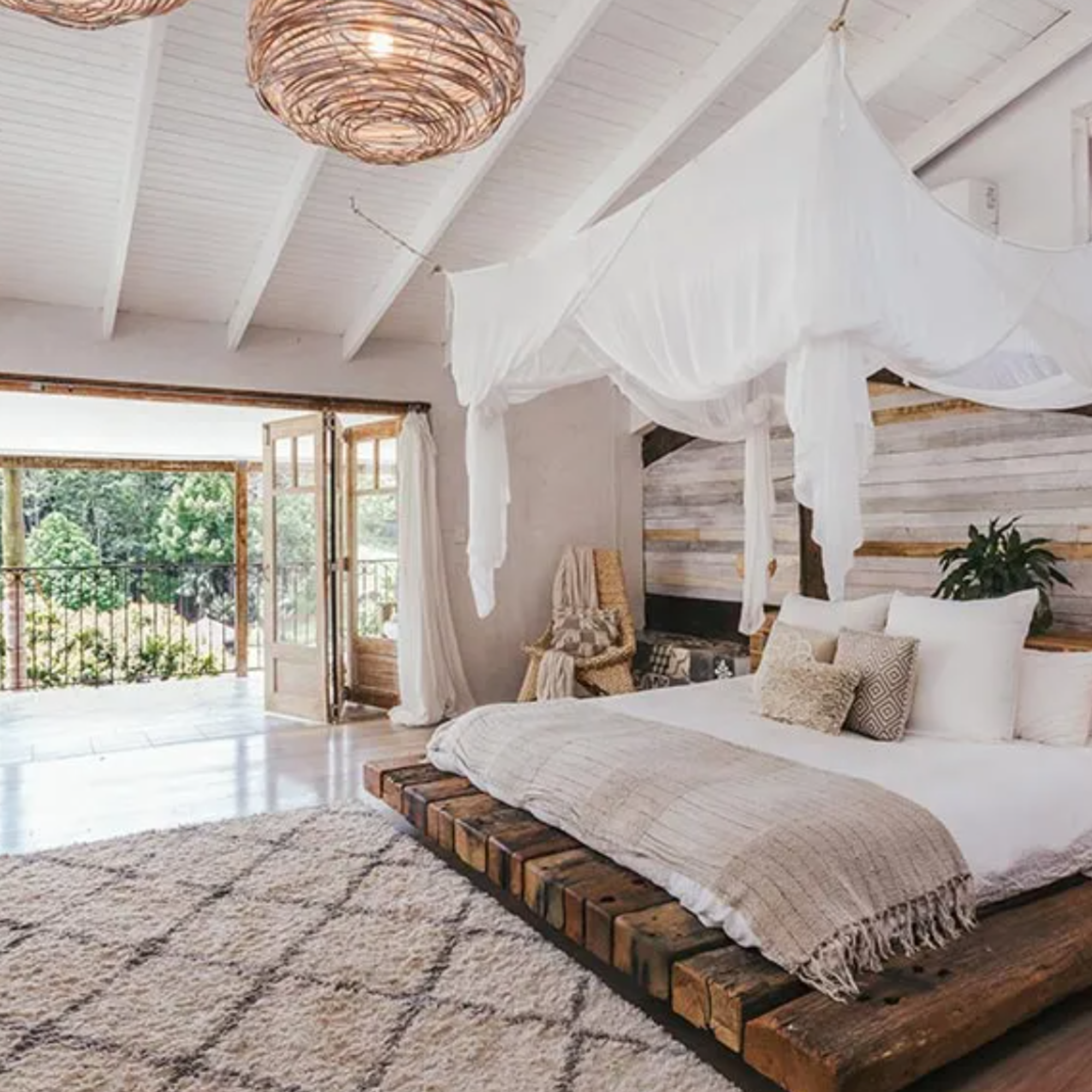 A bedroom with a wooden bed frame, white bedding, and decorative pillows, with a canopy above the bed, a plush rug, and large windows opening to a balcony with trees outside.