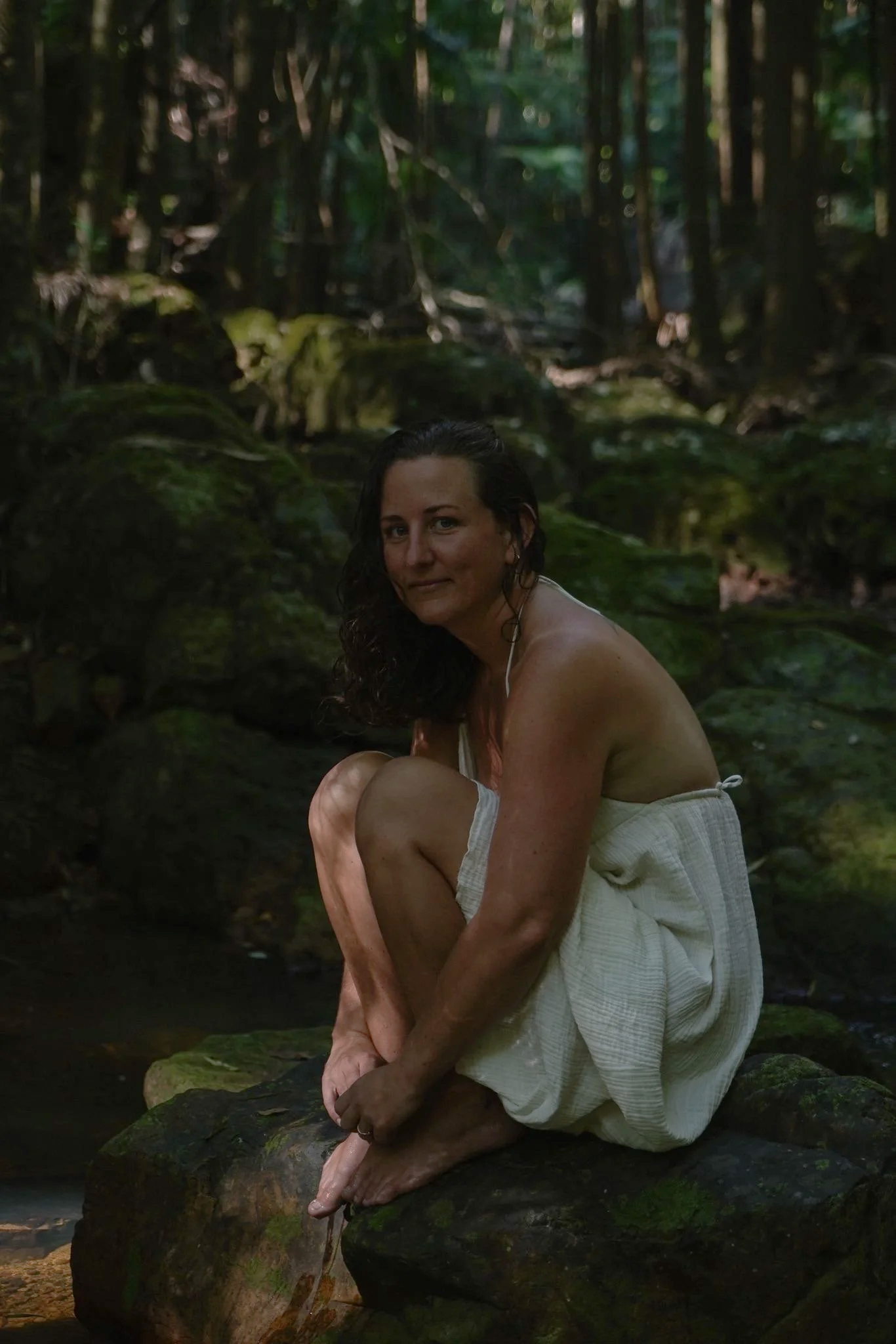 A woman with wet hair sitting on a moss-covered rock in a forest, wearing a white towel, surrounded by green foliage and rocks.