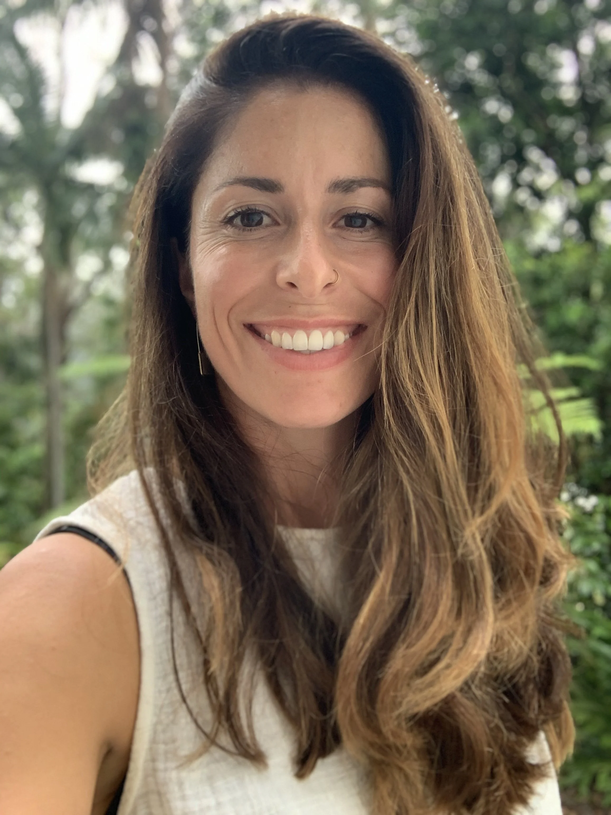 A young woman with long, wavy brown hair taking a selfie outdoors with green trees in the background, smiling with white teeth, wearing a white sleeveless top, with a small nose ring and subtle makeup.