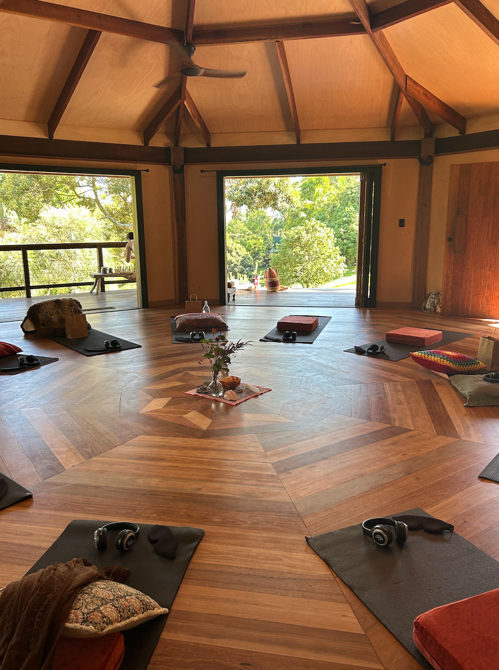 A cozy indoor space set up for a yoga or meditation class with mats, pillows, and headphones, overlooking a lush green outdoor view through open sliding doors.