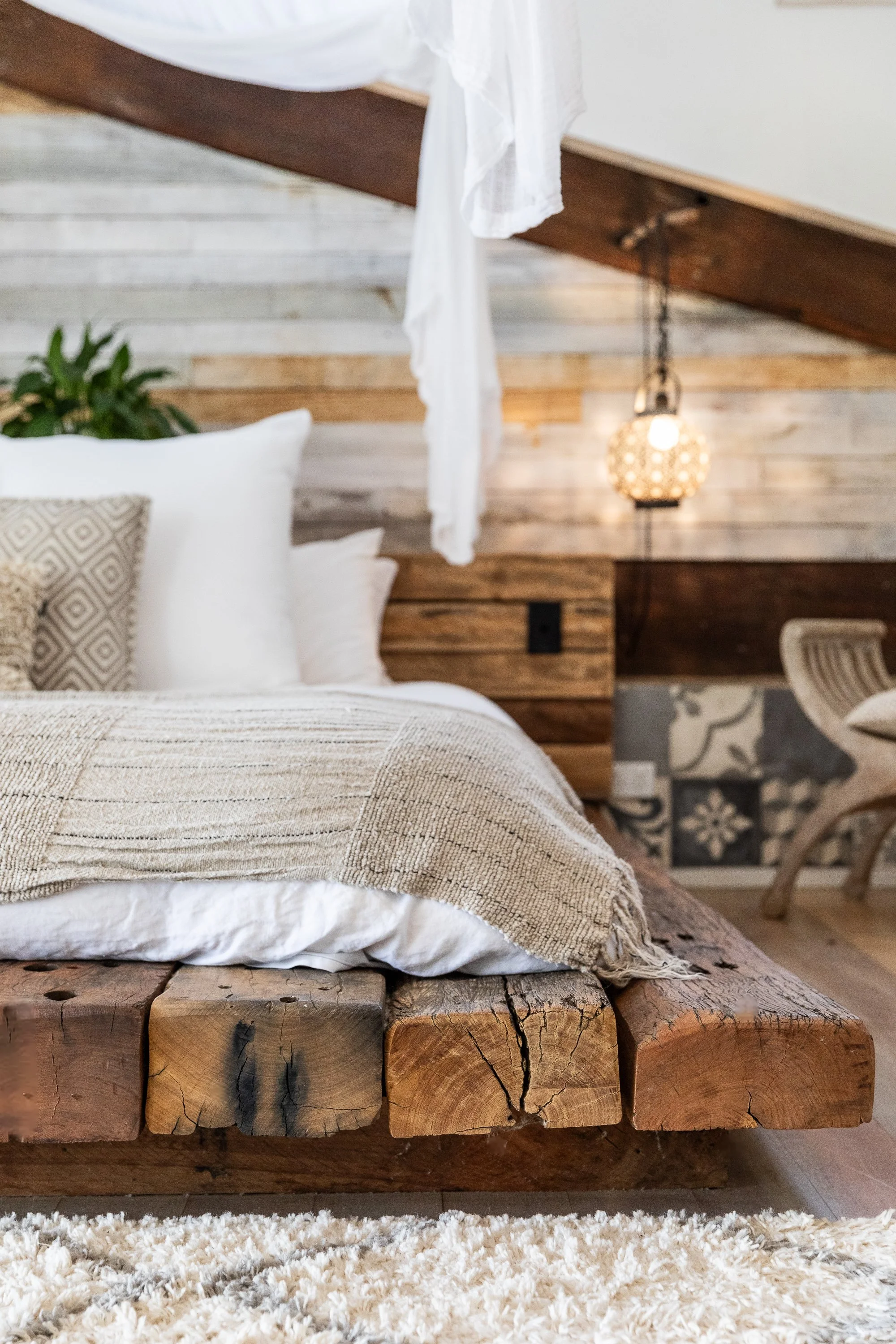 Close-up of a rustic bed with a thick wooden frame, white bedding, and decorative pillows in a cozy bedroom with wooden and patterned walls, a small chair, and a hanging lamp.