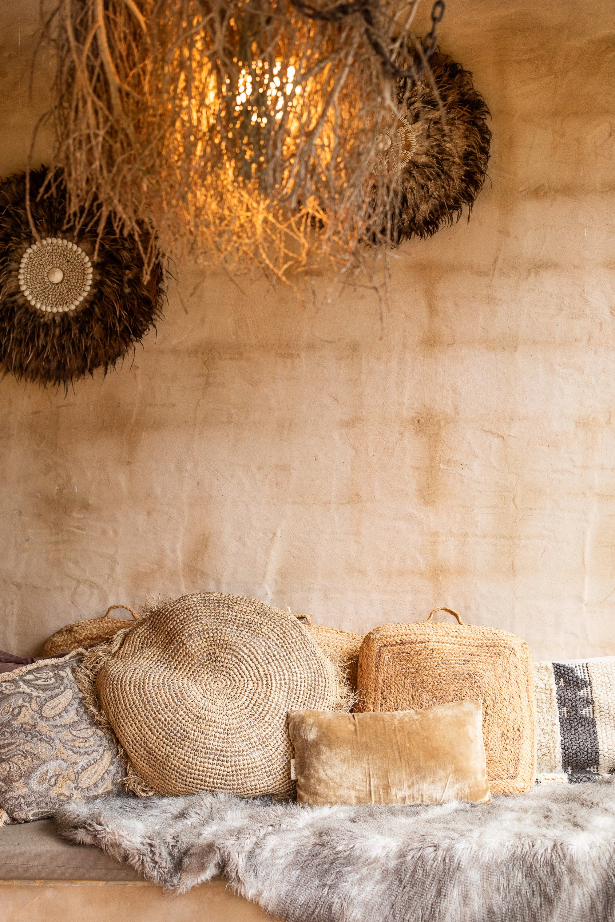 A cozy interior with a beige textured wall, decorated with hanging feather art, and several cushions made of woven materials and velvet, placed on a fluffy faux fur throw on a sofa.