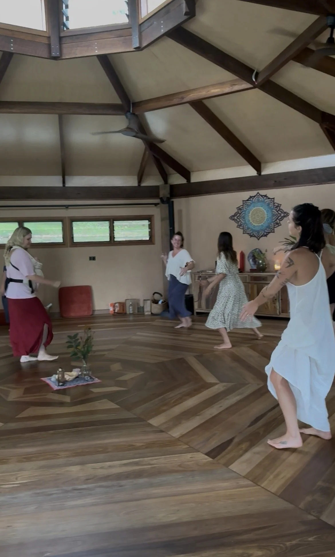 Group of women dancing or practicing yoga together in a spacious room with wooden floors and high vaulted ceiling.