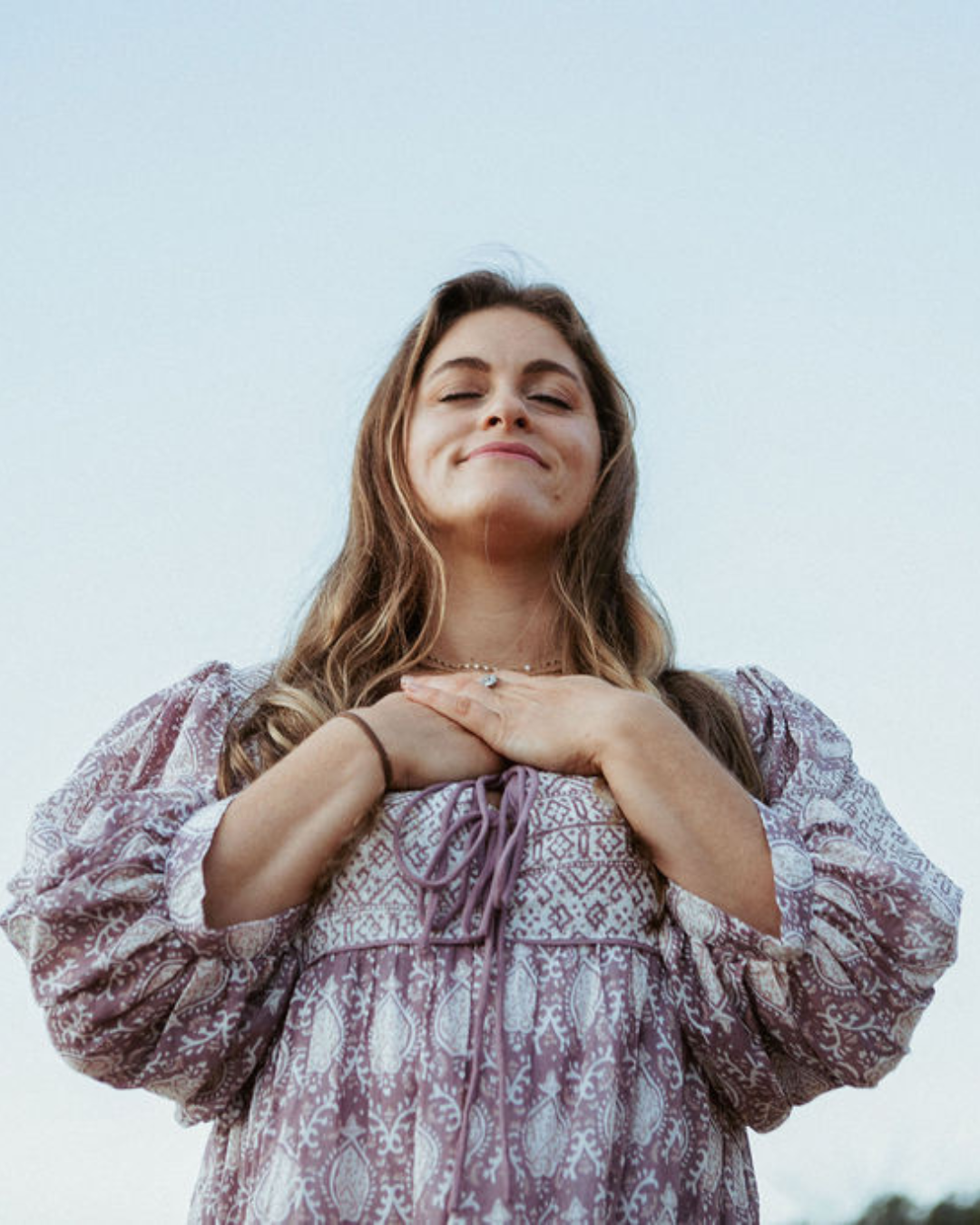A woman with long, wavy hair standing outdoors with her eyes closed and hands crossed on her chest, smiling in front of a clear sky.