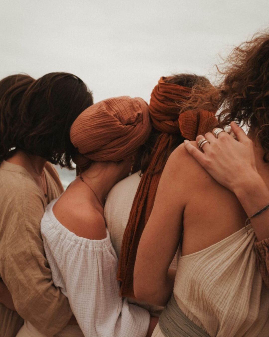 A group of women hugging with their heads bowed and arms wrapped around each other, wearing earth-toned clothing and headscarves.
