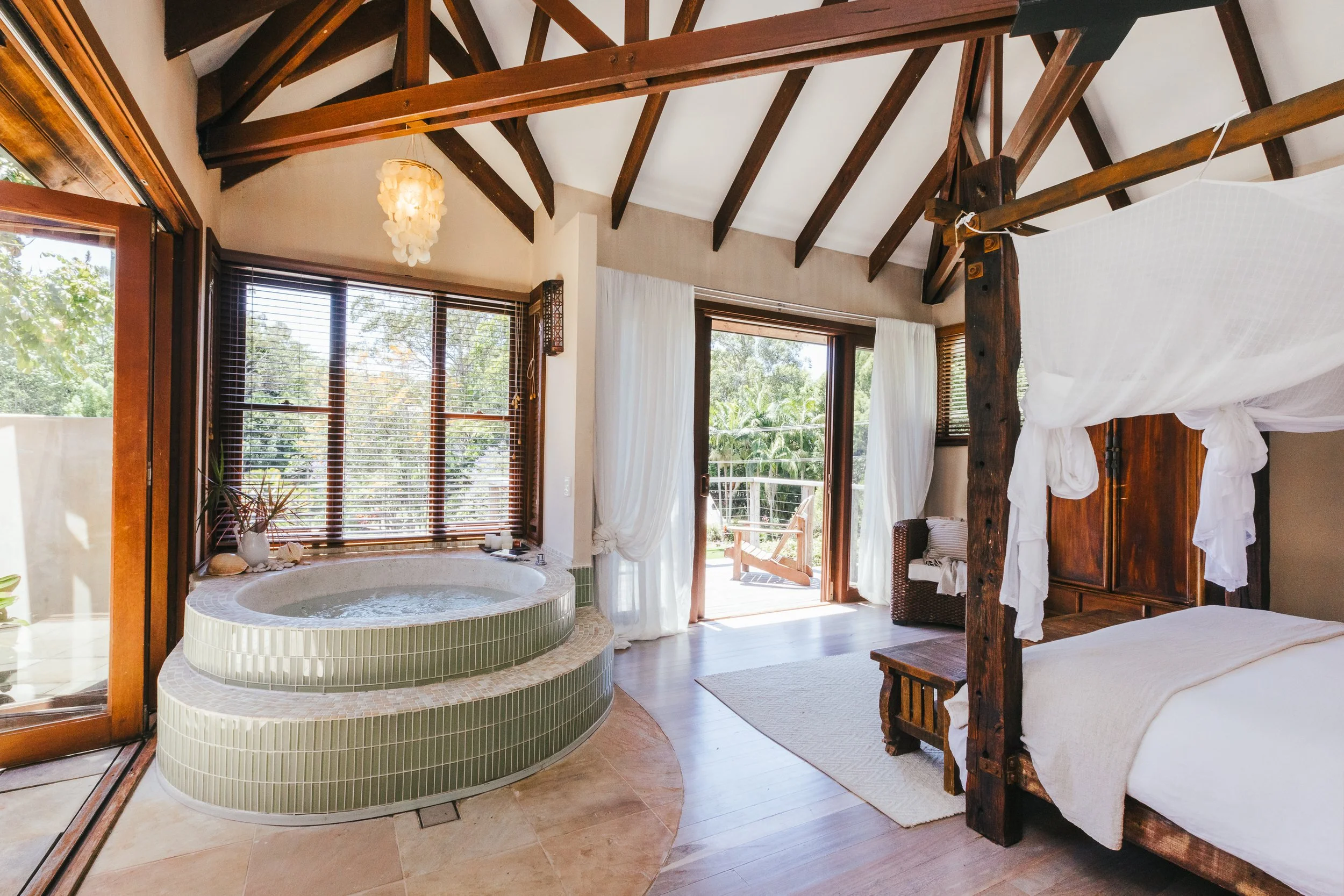 A bedroom with a wooden four-poster bed with white bedding, a small sitting area with a chair and cushions, large windows with blinds, an open sliding door leading to an outside deck with lounge chairs, a jacuzzi with green tiles, a potted plant, and a wooden ceiling with exposed beams.