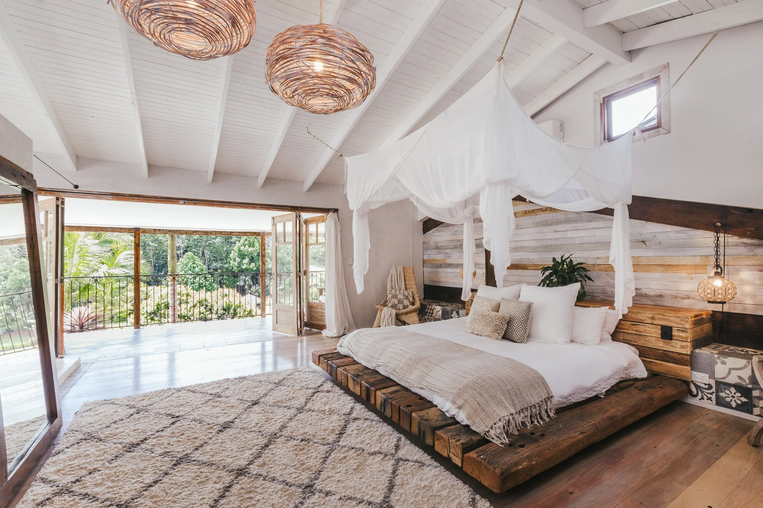 A bedroom with a rustic wooden bed and a white canopy, pillows, and a beige throw blanket, with a view of a balcony with open doors and greenery outside.