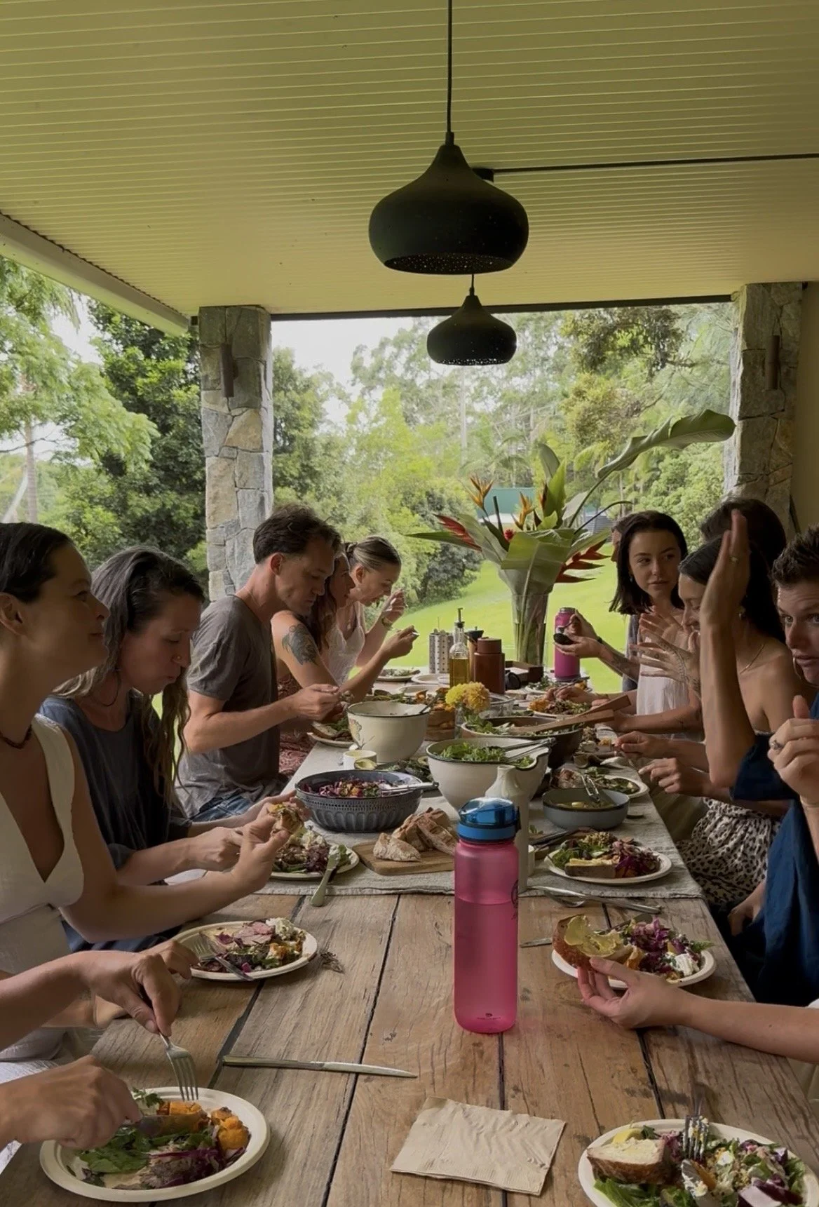 People dining together at a long outdoor table with various food and drinks, overlooking a lush green landscape.