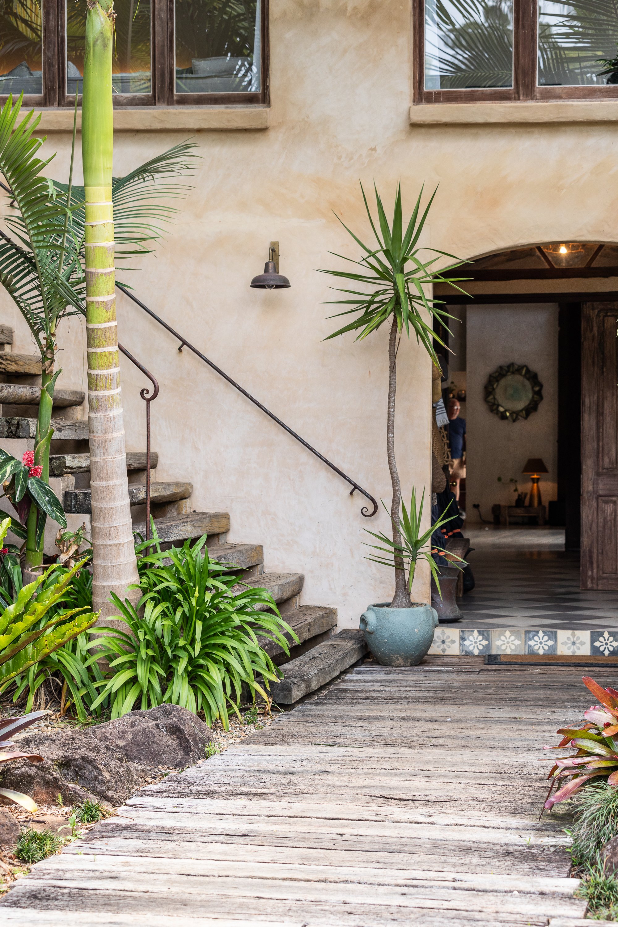Exterior scene of a house with tropical plants, wooden stairs, and a wooden plank pathway leading to an open entryway.