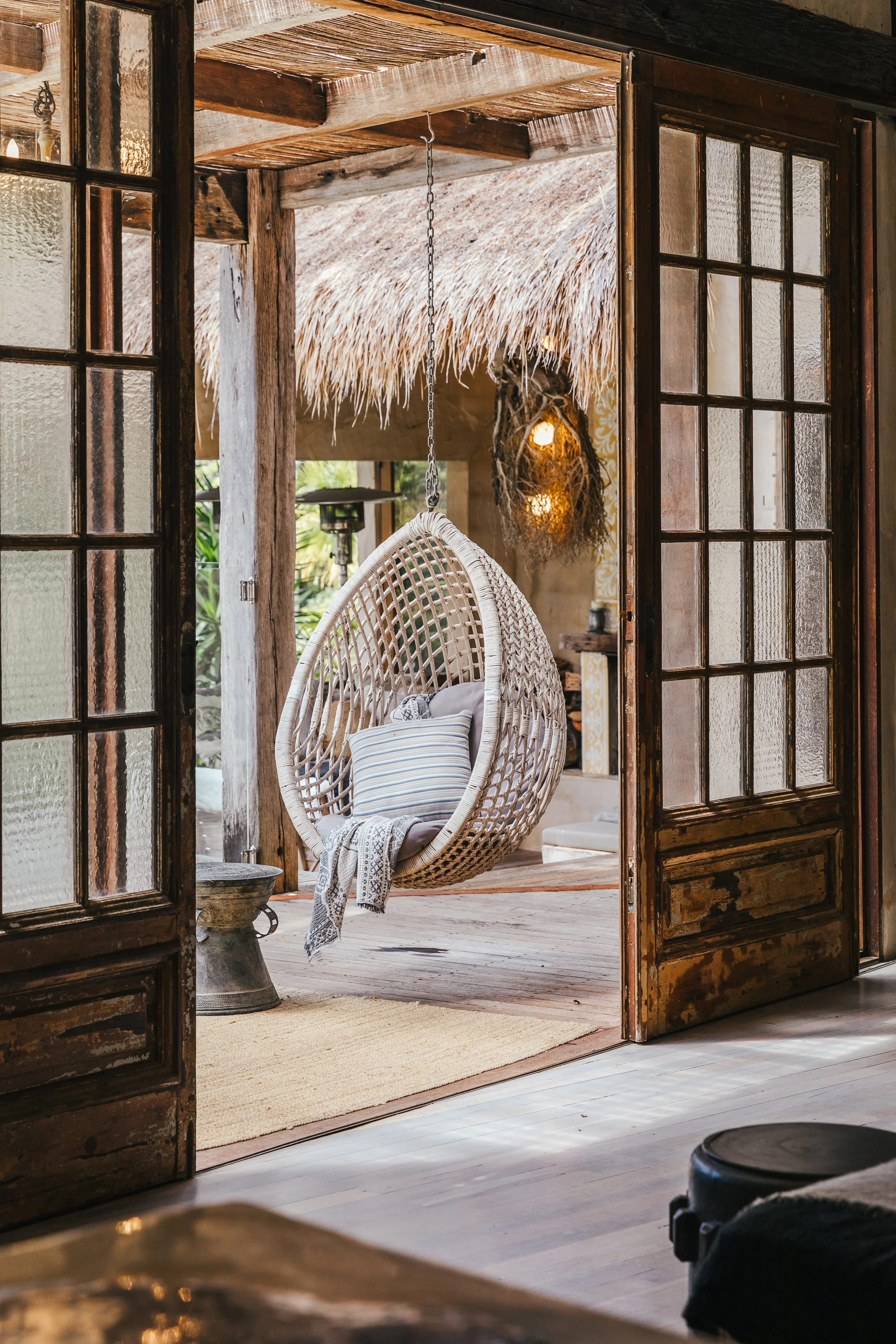 A cozy outdoor hanging egg chair with pillows and a blanket in a tropical-style space, visible through open wooden and glass sliding doors.