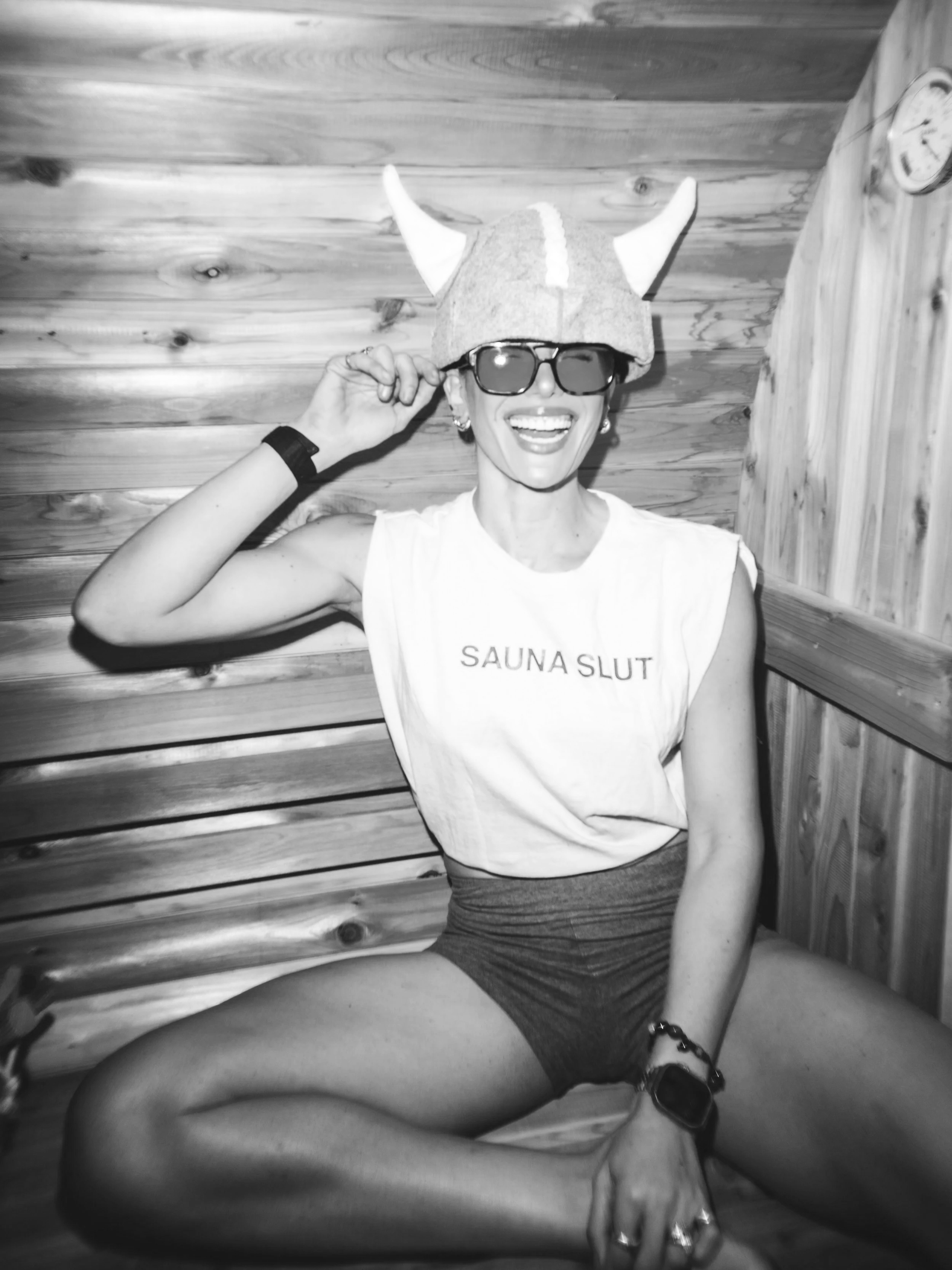 Nikki Howard is sitting in a wooden sauna, wearing shop sauna slut wellness merchandise.