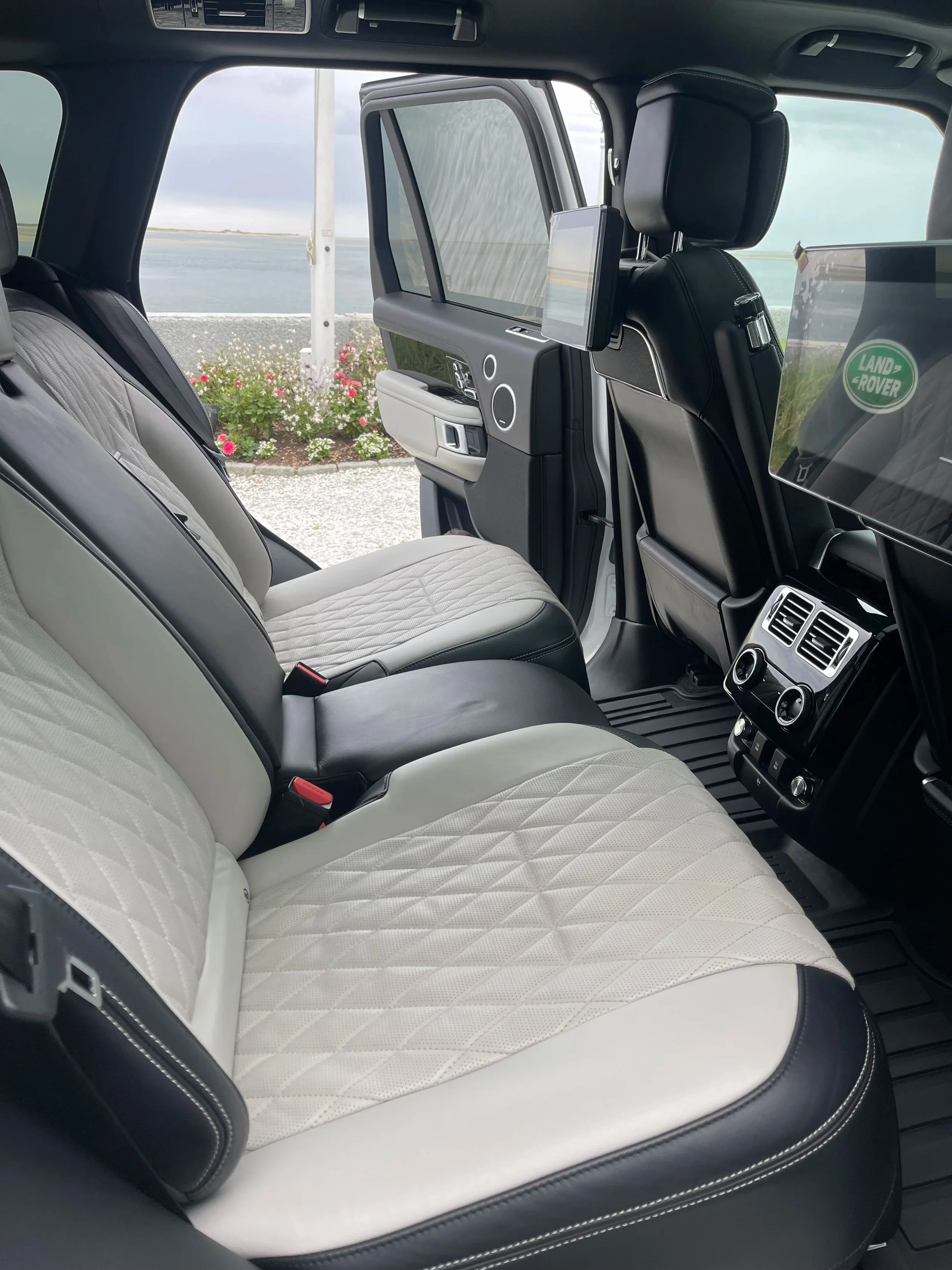 Interior of a luxury vehicle with white and black quilted seats, a view of the water and flowers outside through the open door, and screens mounted on the back of the front seats.