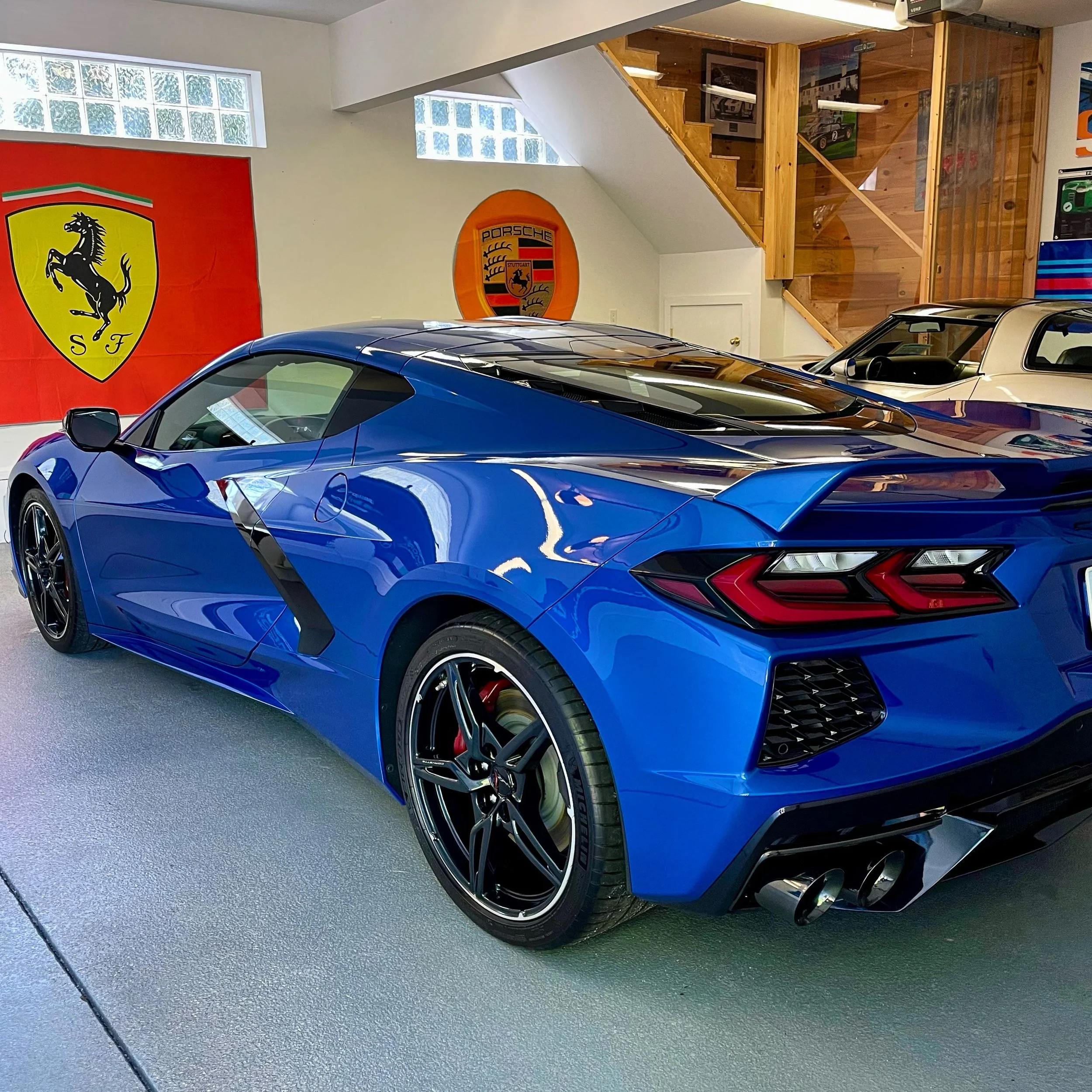 Blue sports car parked indoors with Ferrari and Porsche logos on the wall.