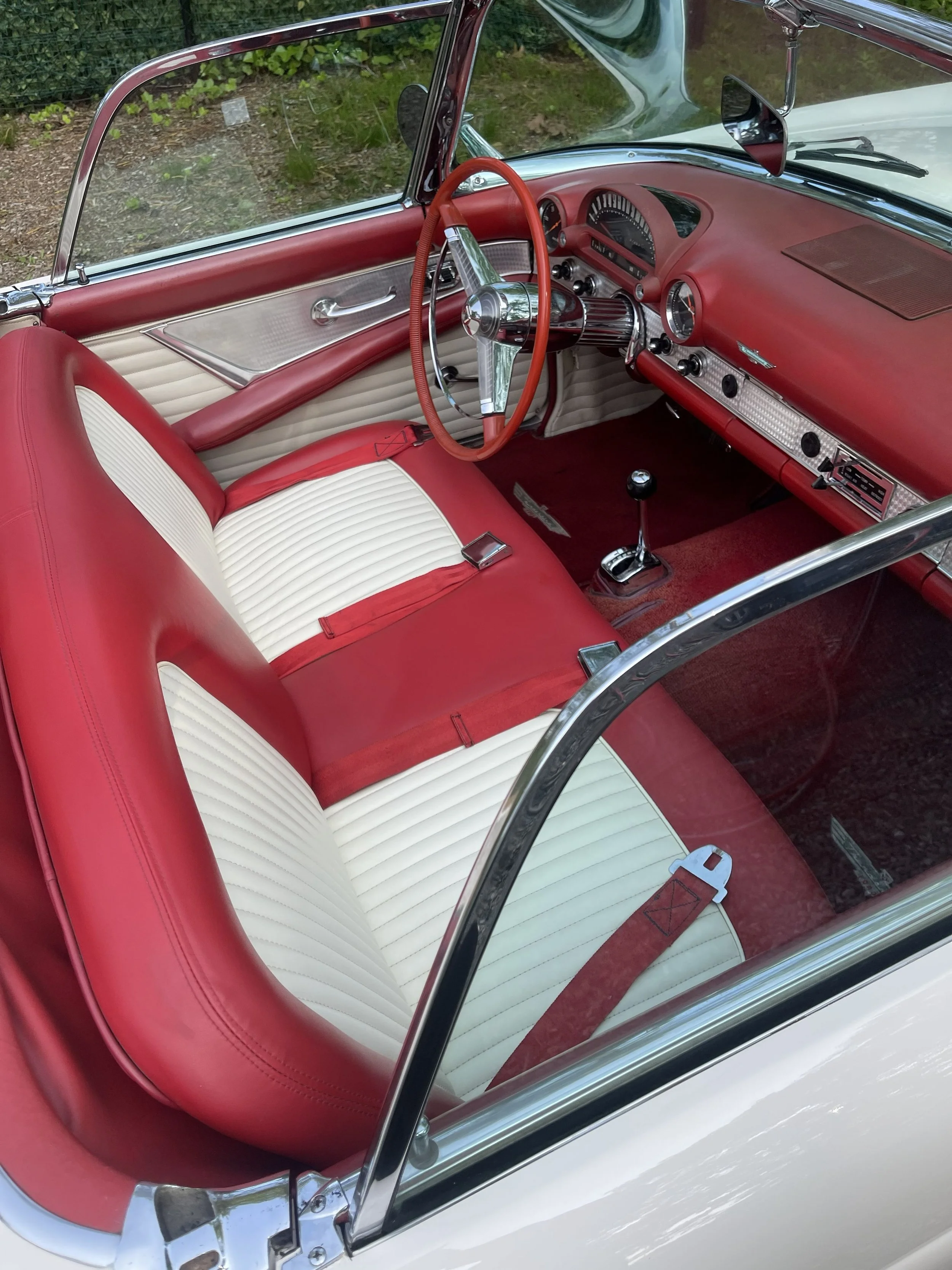 The interior of a vintage car with red and white seats, a red dashboard, and a red steering wheel, viewed through the windshield.