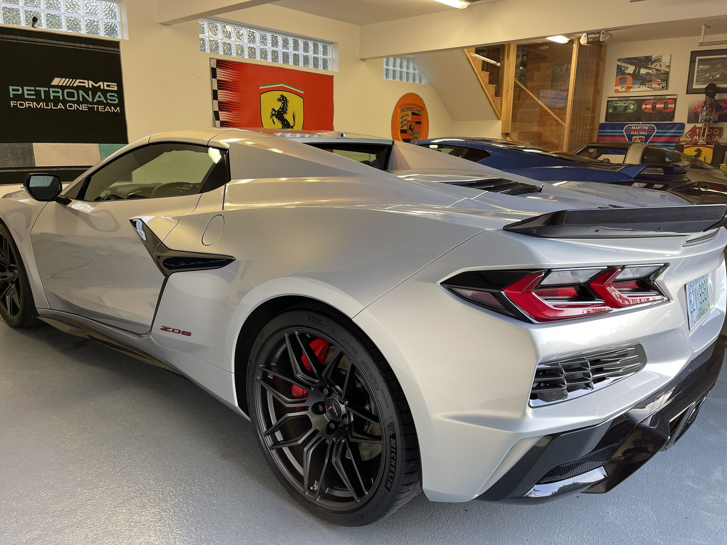A white Lamborghini sports car with black and red wheels parked indoors, surrounded by racing and luxury car posters and banners on the walls.
